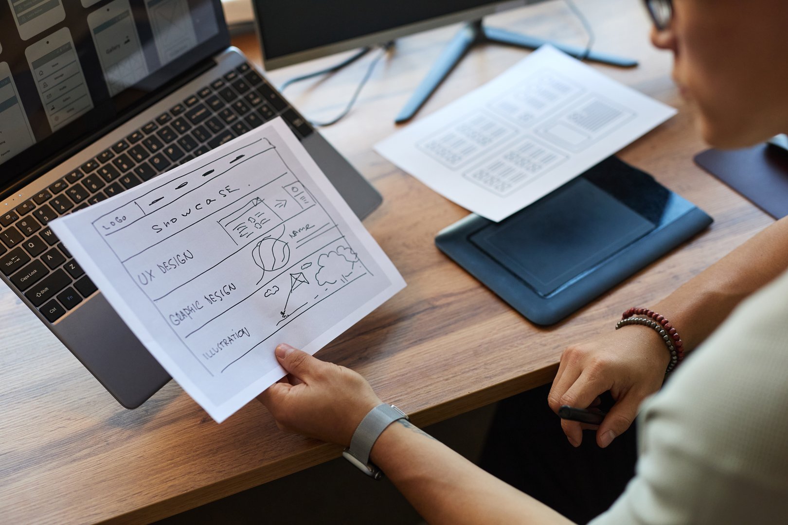 Young adult man reviewing website wireframe sketch while sitting at desk with laptop and graphics tablet, holding printed design concept and analyzing project layout