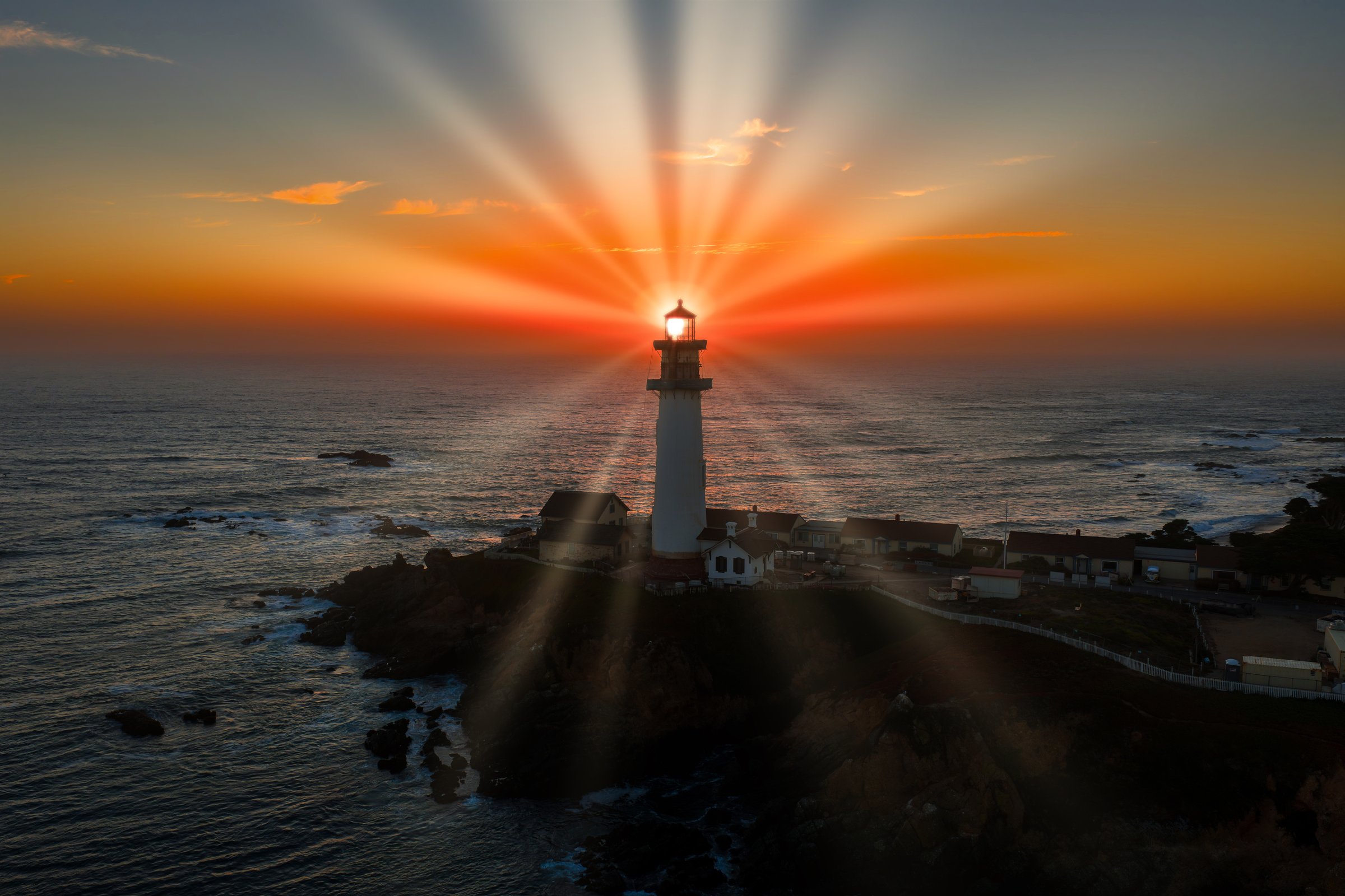 Pigeon Point Lighthouse stands illuminated at sunset, casting light beams across the sky. The rocky shoreline and Pacific Ocean complete the coastal scene.