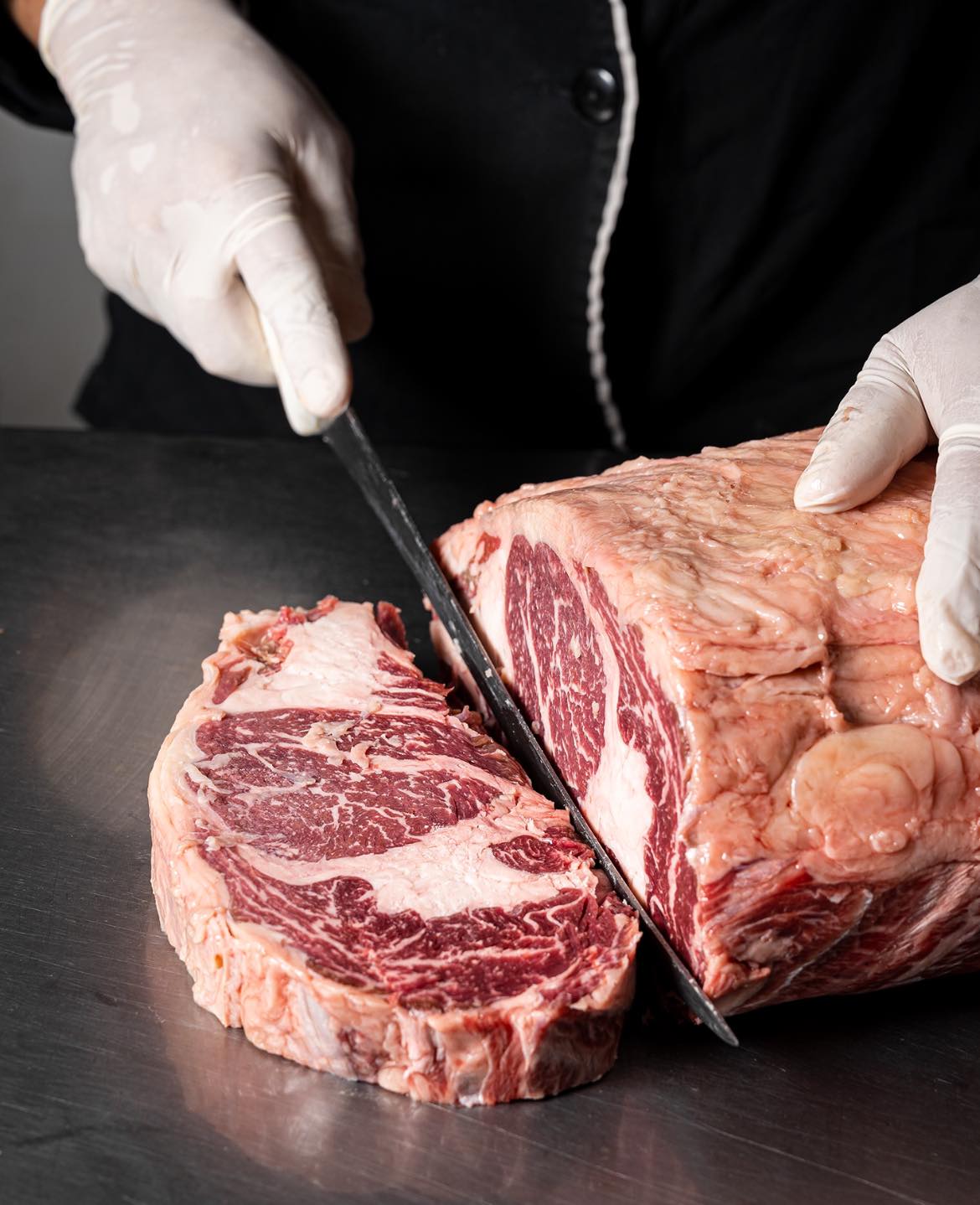 Person slicing raw ribeye steak with marbled fat on a cutting board, wearing gloves and using a large knife.