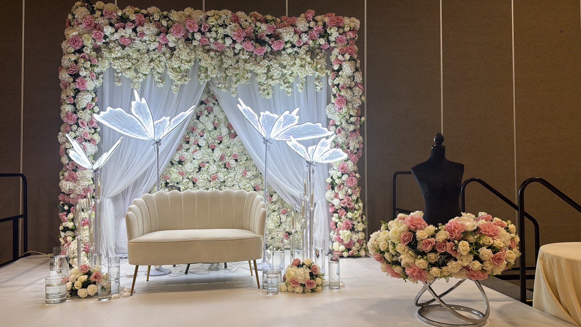 Elegant wedding stage with white couch, floral backdrop, and mannequin adorned with pink and white flowers.