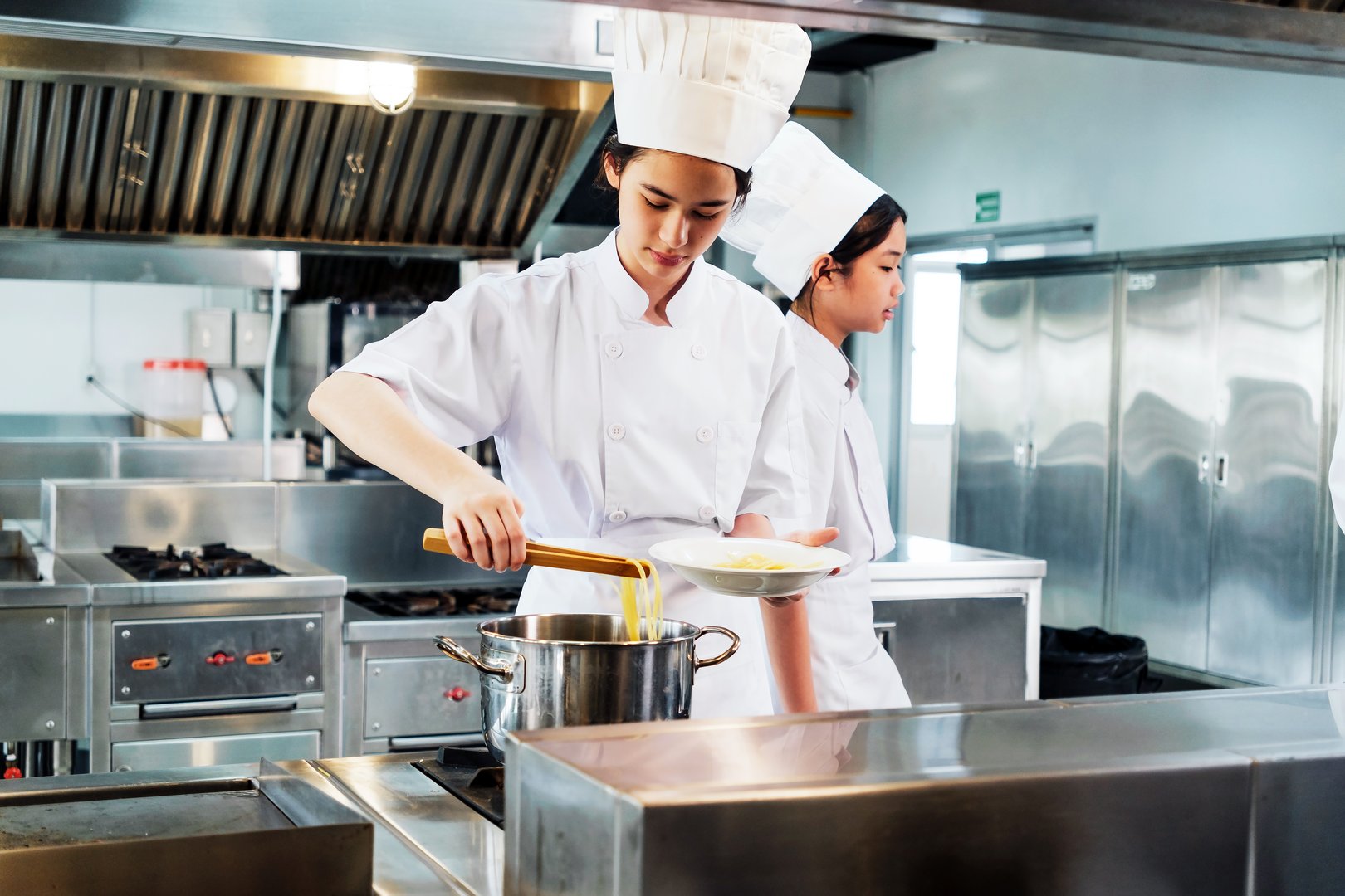 Young culinary student in a white chef uniform plating hot pasta from a steaming pot in a professional kitchen, demonstrating practical cooking skills during chef training at a culinary institute.