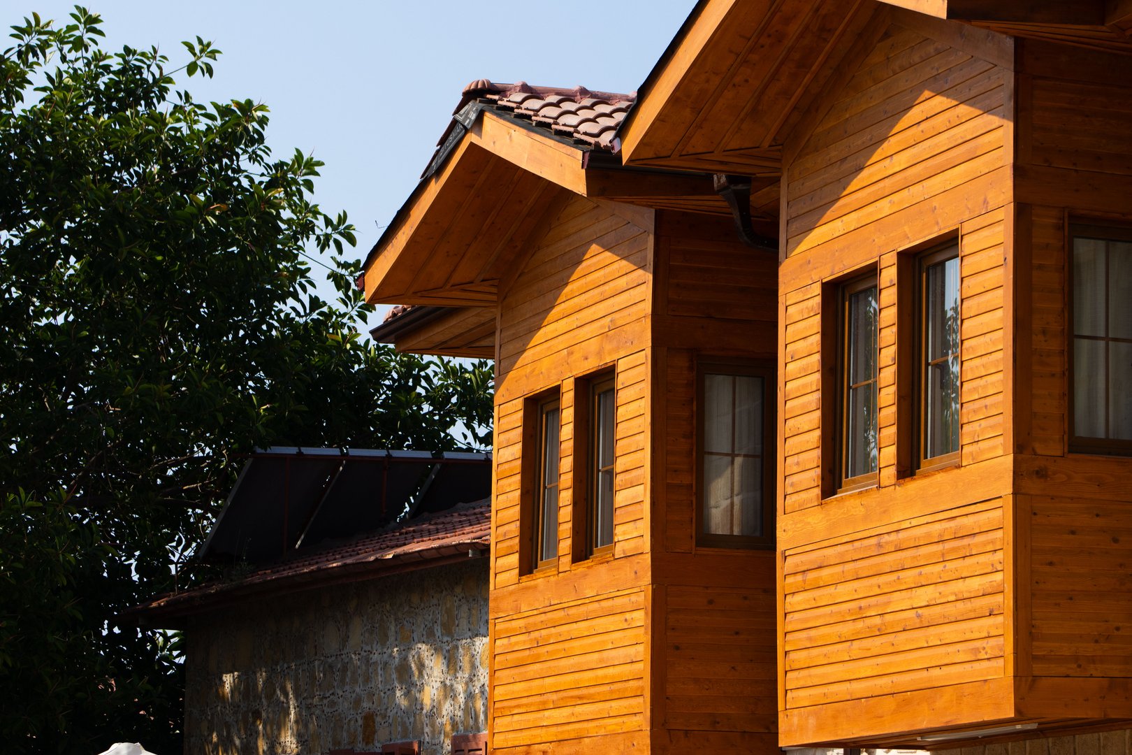 Two warm-toned wooden houses with contemporary lines are seen alongside a traditional stone building with a terracotta roof, showcasing a blend of architectural styles common in the region