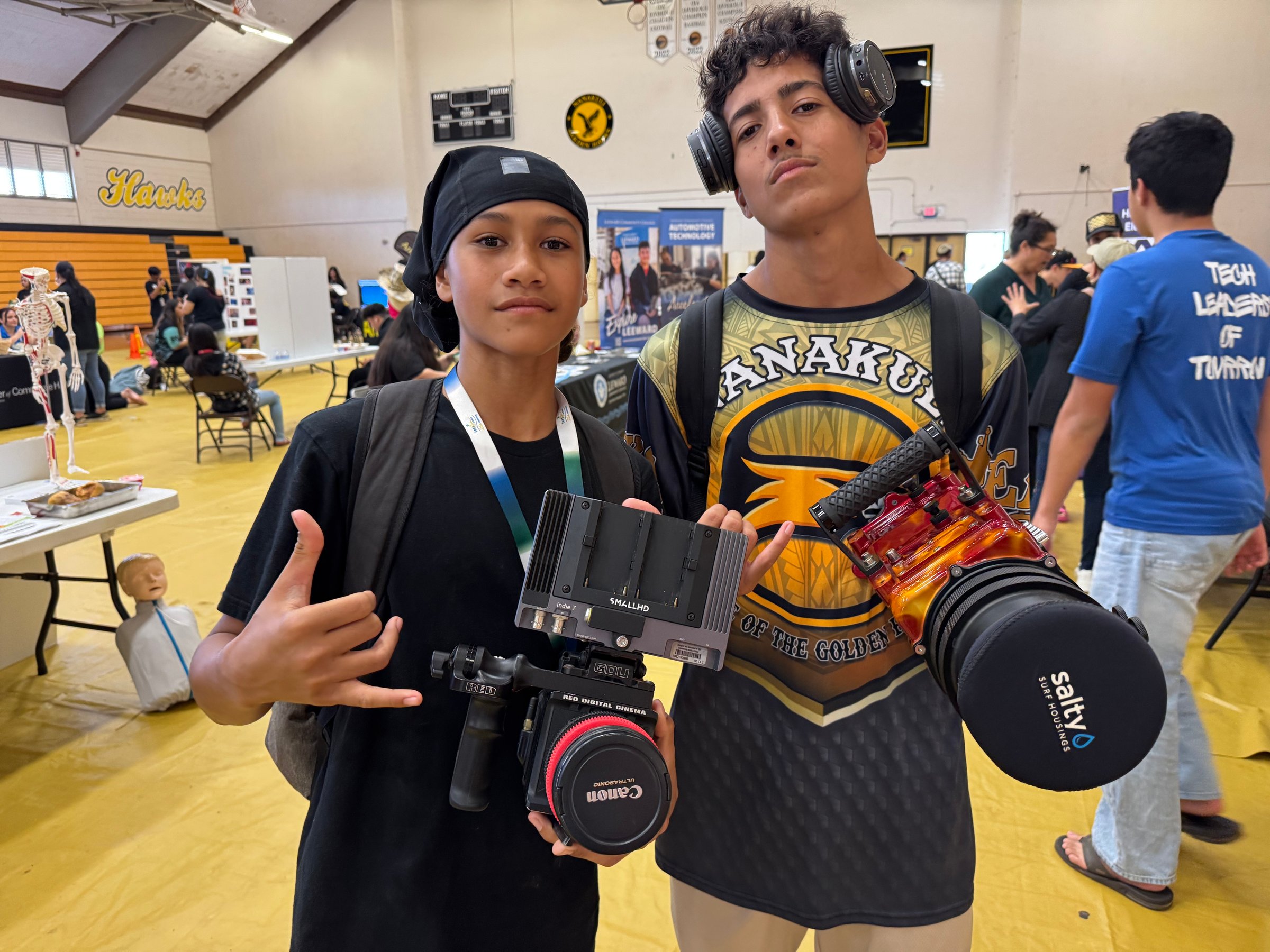 Two young people holding professional cameras in a gymnasium with booths and tables in the background.