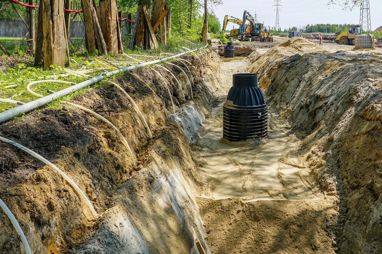 Trench dewatering system installation on a construction site, showing exposed pipes, sand bedding, and new black plastic underground utilities well