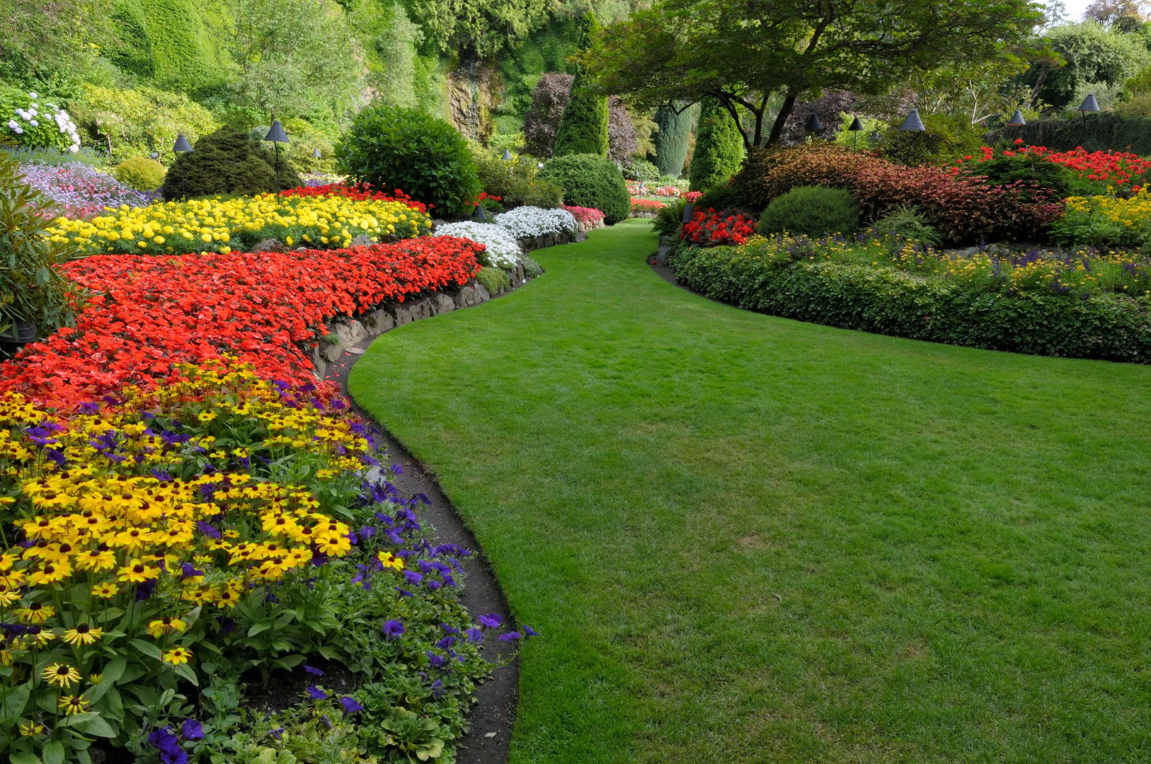 A lush garden with vibrant flower beds, featuring red, yellow, purple blooms, and a winding green lawn path.