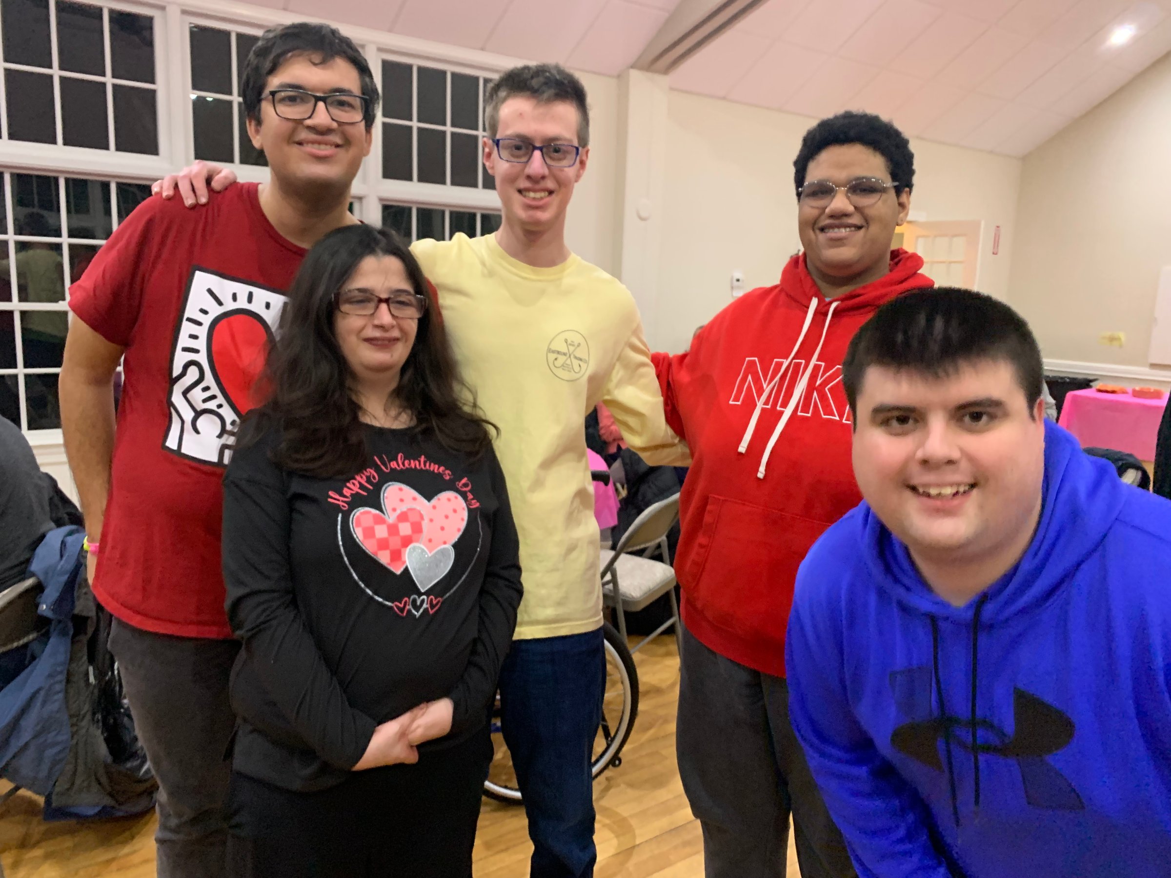 Five people smiling and posing together indoors; three standing, one sitting in a wheelchair, and one kneeling.