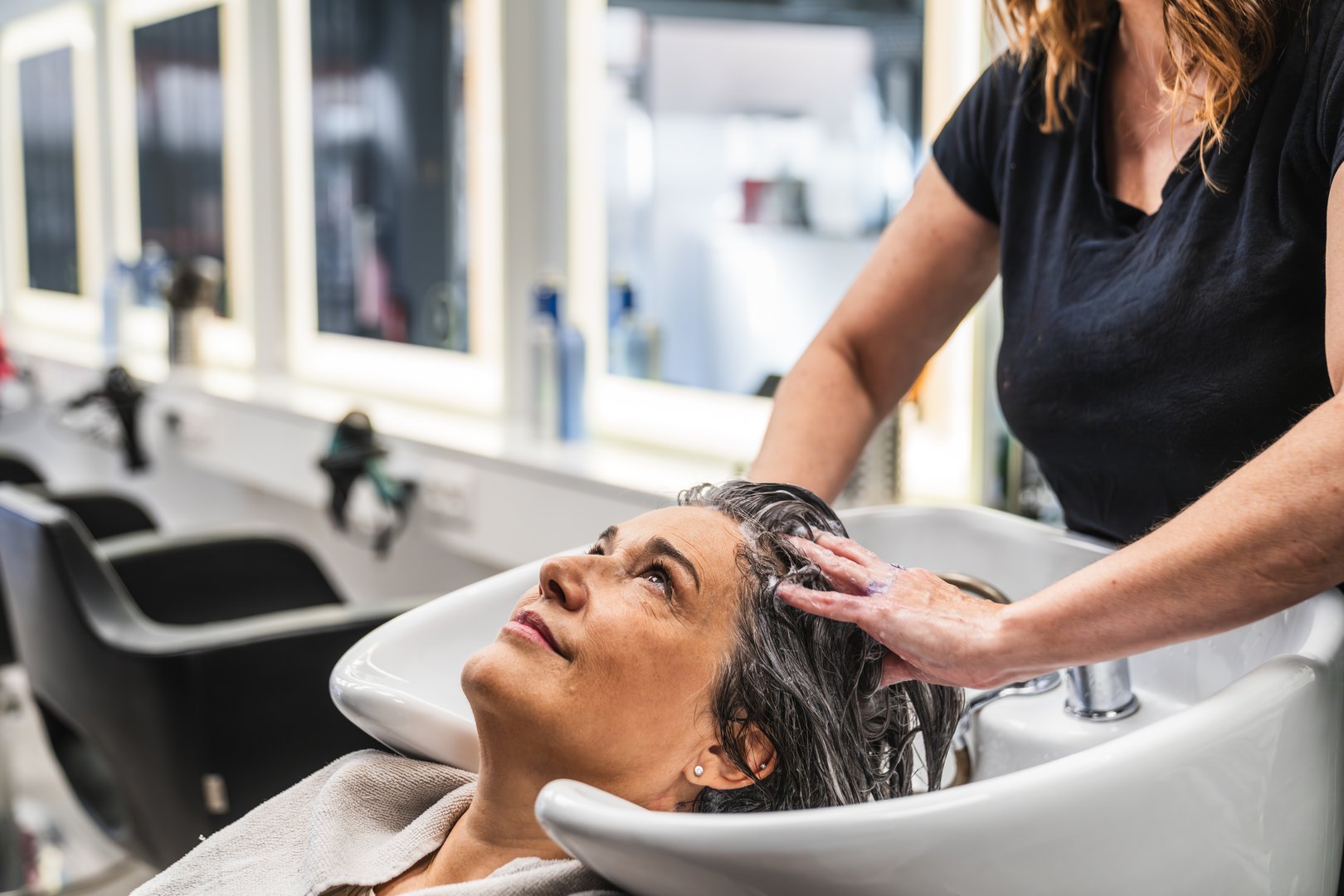 Mid adult woman enjoying a relaxing shampoo and head massage by a professional hairdresser at a modern, bright salon sink