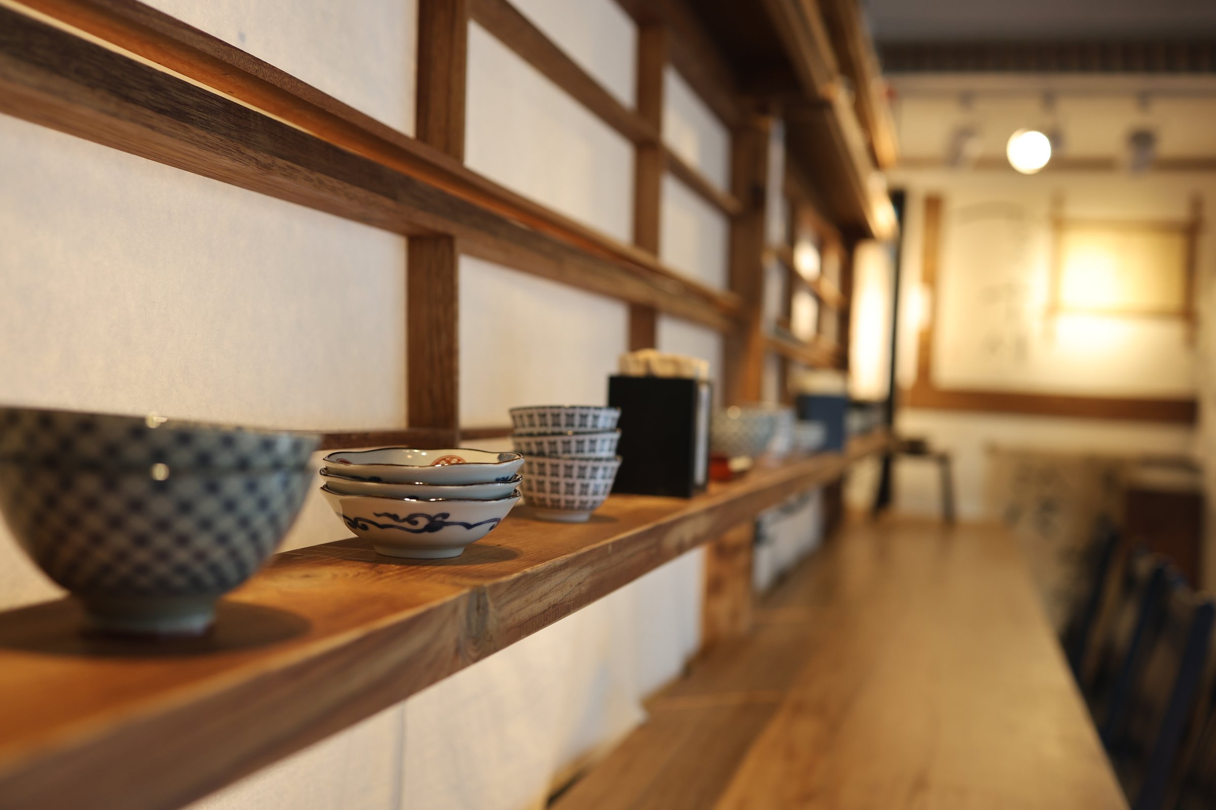 a serene and neatly arranged display of traditional Japanese tableware on wooden shelves, featuring a variety of bowls and containers, each with unique patterns and designs. The warm lighting and soft focus create an inviting atmosphere, reflecting the artistry and cultural significance of the items in a Japanese restaurant setting.
