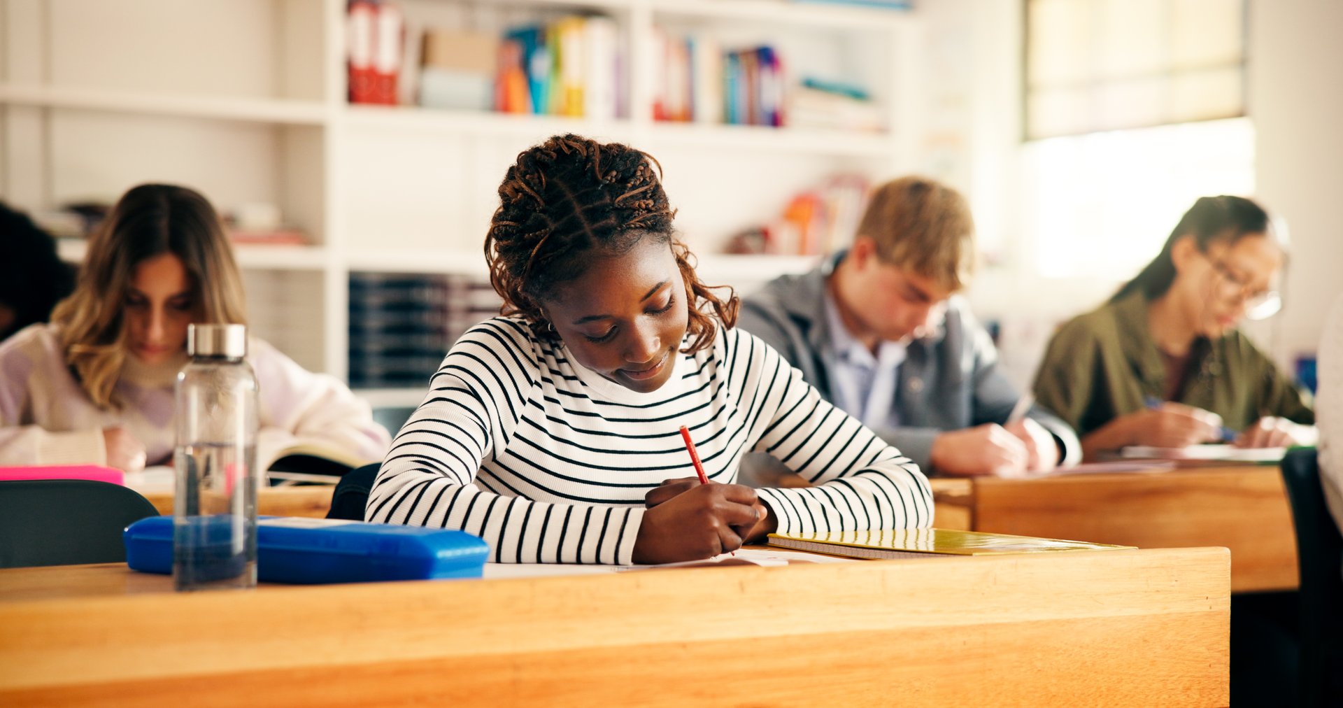 High school students, girl and writing in lesson for assessment quiz, evaluation test or learning. Teenager, education and people with exam essay in classroom for scholarship or curriculum knowledge