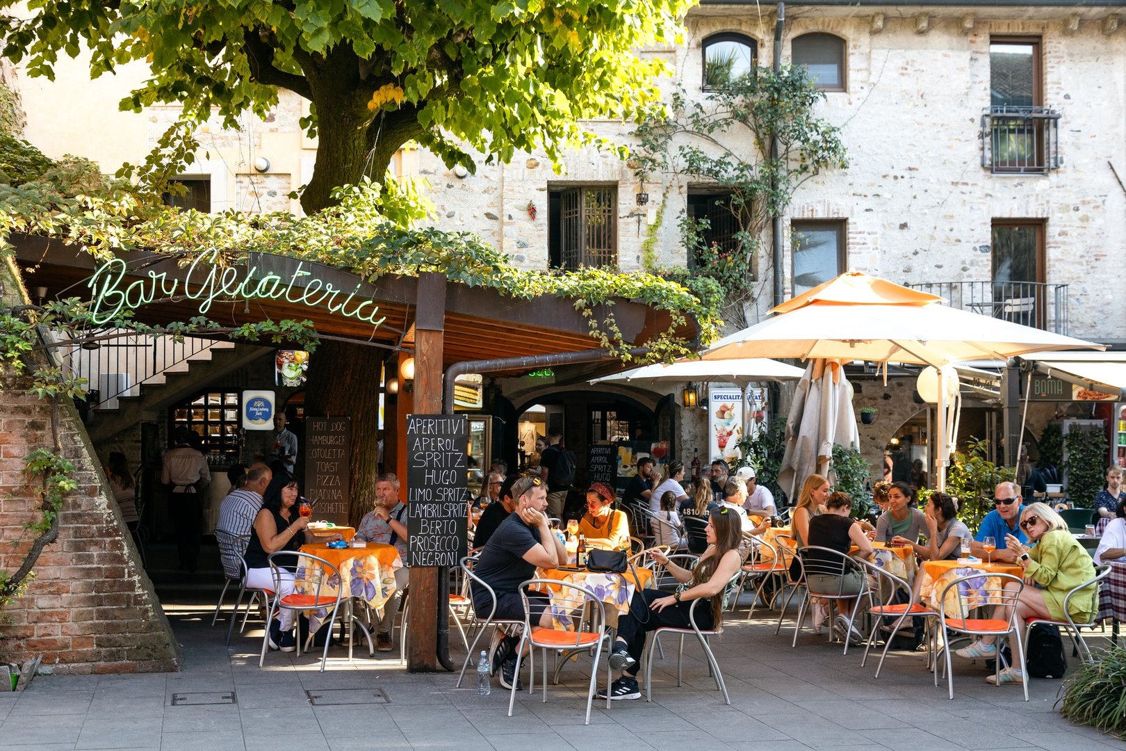 Sirmione, Italy - Sep 21st, 2024: People enjoying aperitivo at a cozy outdoor gelateria under the shade of trees