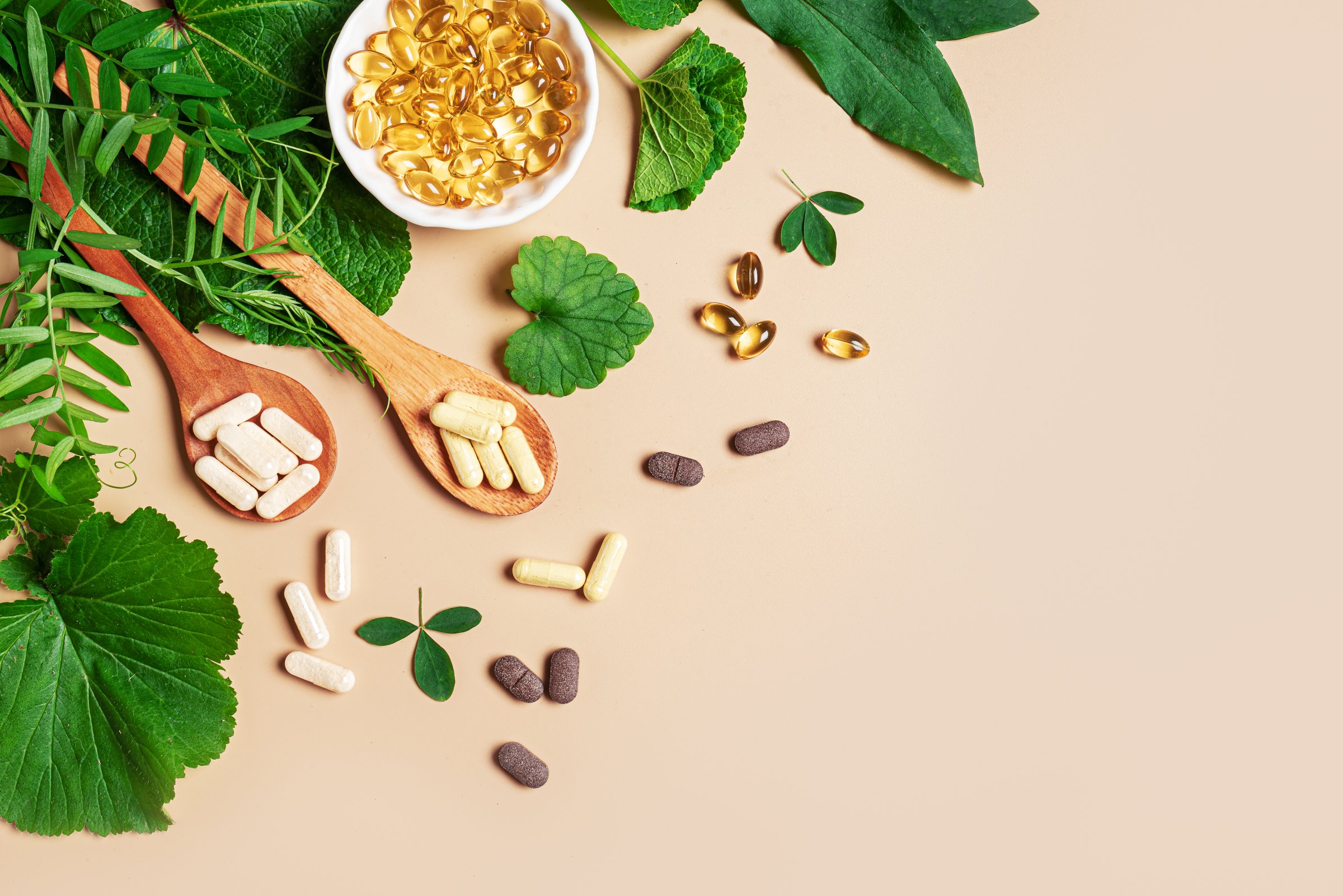Plant based supplements. Organic vitamins. Homeopathy and alternative herbal medicine. Vegan capsules and pills, green plant leaves on beige background, top view, copy space.