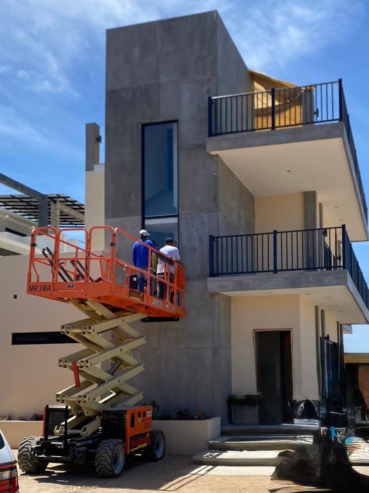 Two workers on a scissor lift inspecting a modern two-story house with balconies and large windows.