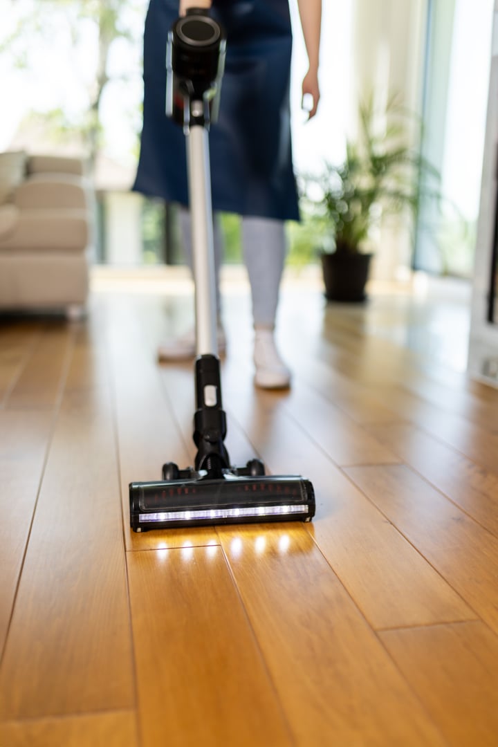 A person is using a vacuum cleaner on hardwood flooring in a bright, airy living room. The sunlight filters through large windows, adding warmth to the clean environment.