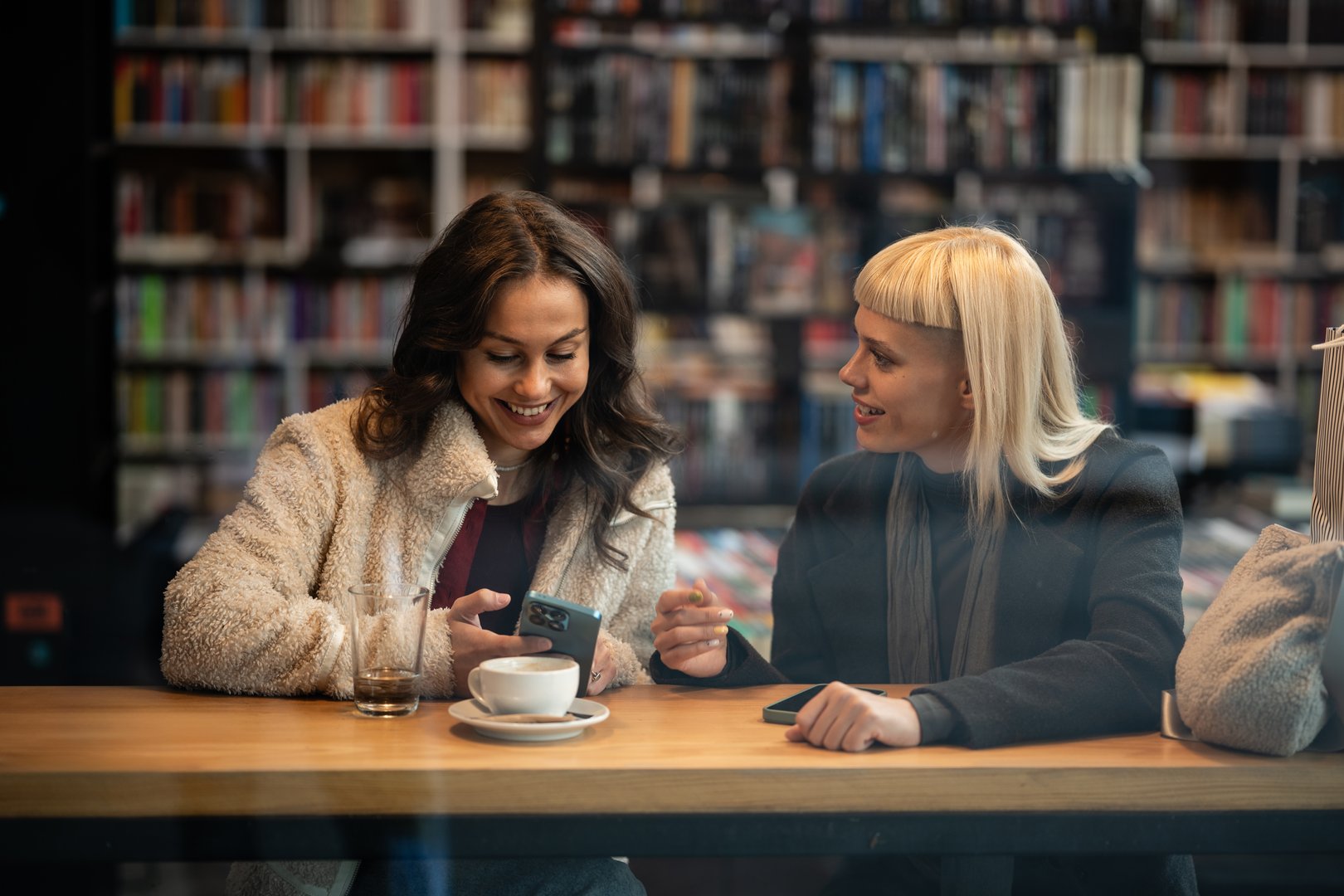 Two young women friends enjoying coffee and conversation, sharing content on a smartphone in a cozy library cafe