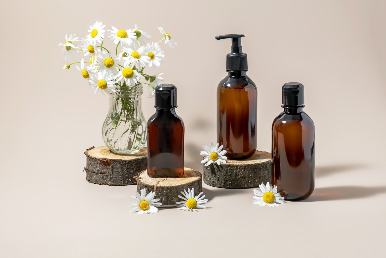 Dark brown bottles of cosmetics on wooden catwalks and a vase with chamomile flowers on a beige background. Packaging design of shampoo bottles for spa baths, shower gel dispenser.