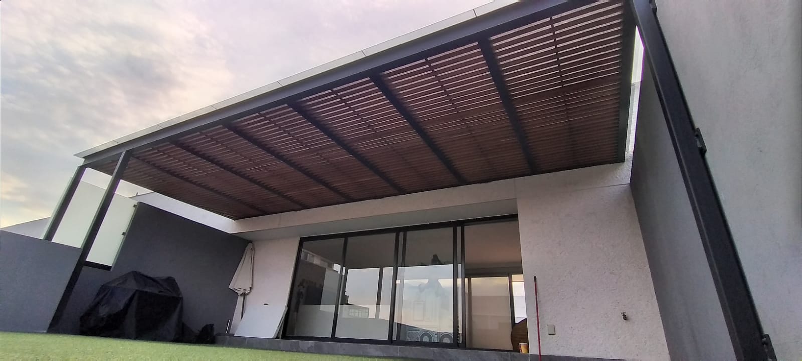 Modern patio with wooden pergola, sliding glass doors, and outdoor seating area under a cloudy sky.
