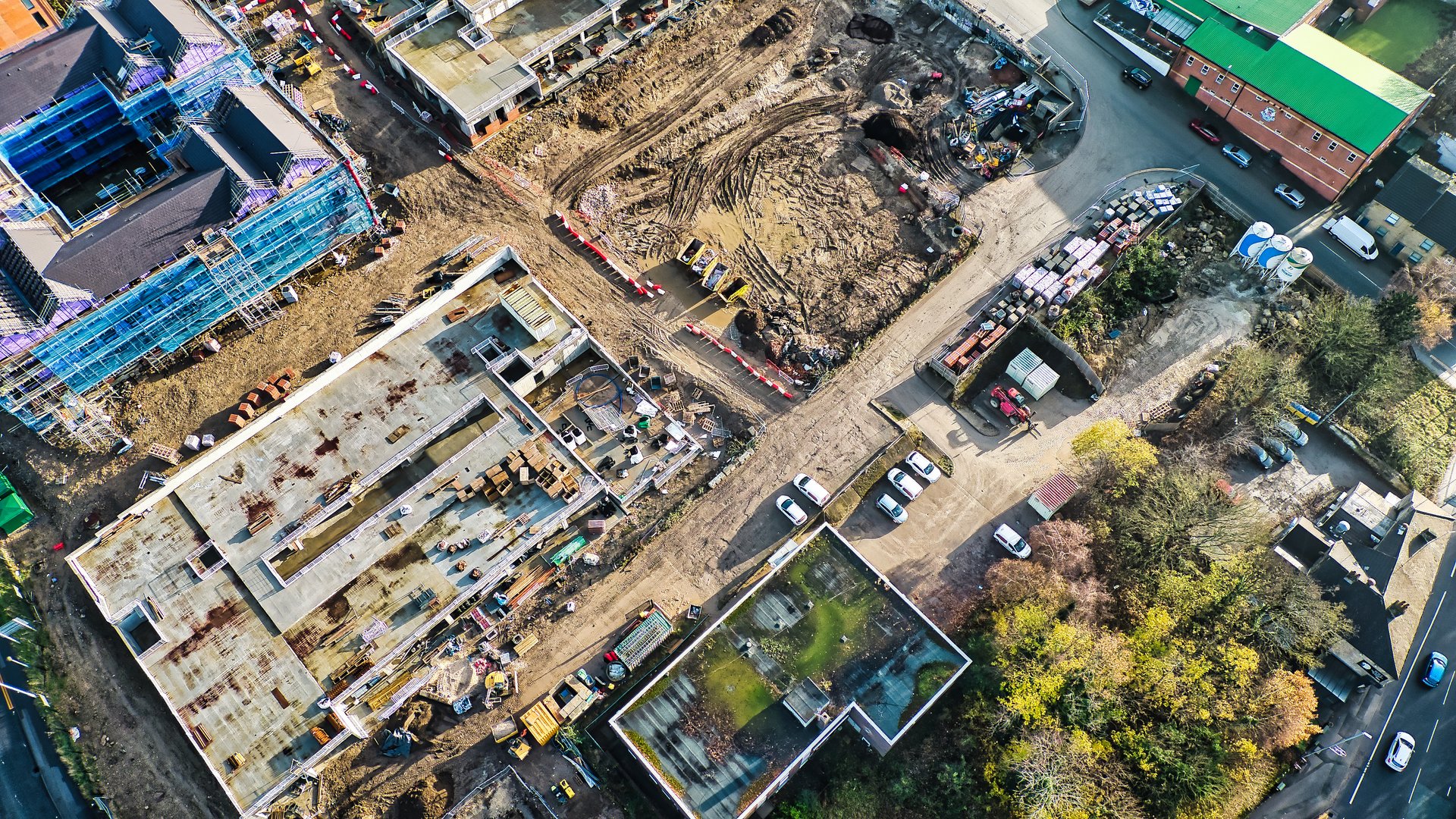 Aerial view of a construction site showing buildings under construction, excavated land, parked vehicles, and surrounding greenery.  Multiple structures in various stages of completion are visible.