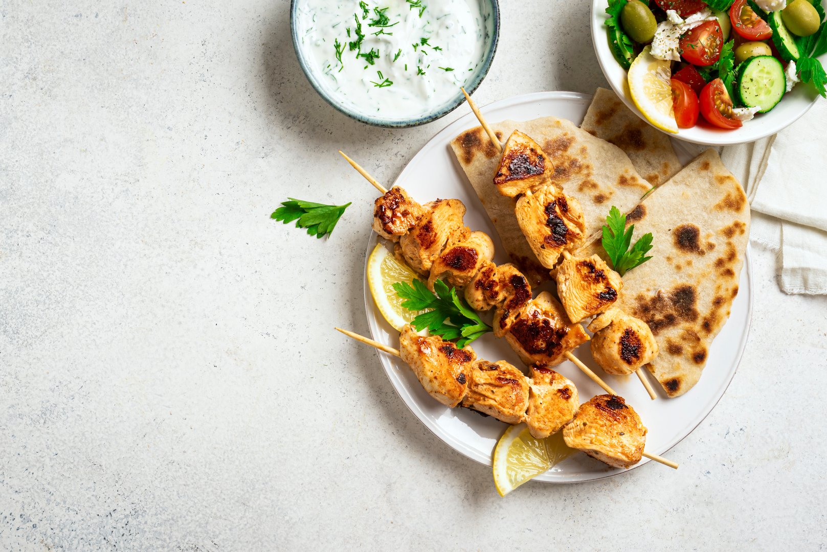 Mediterranenan healthy lunch: chicken skewers souvlaki, flatbread and vegetable salad. Fried chicken kebabs, greek salad, tzatziki yogurt sauce and pita bread on white table, copy space, top view.