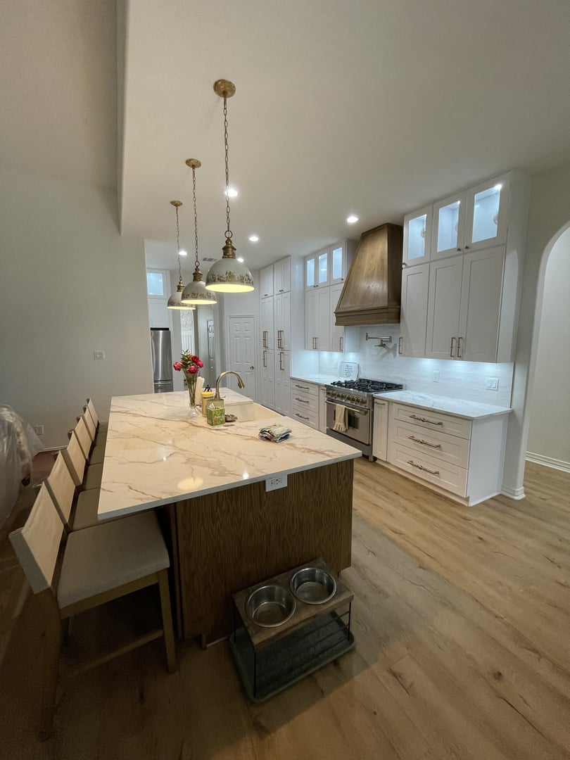 Modern kitchen with white cabinets, marble island, pendant lights, and wooden floor. Stainless steel appliances visible.