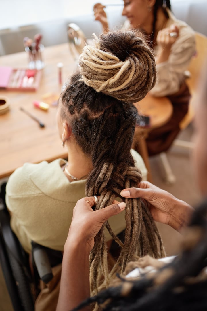 Woman braiding long dreadlocks of girlfriend sitting in wheelchair while another woman with similar hairstyle sitting at table in background, beauty tools visible on tabletop