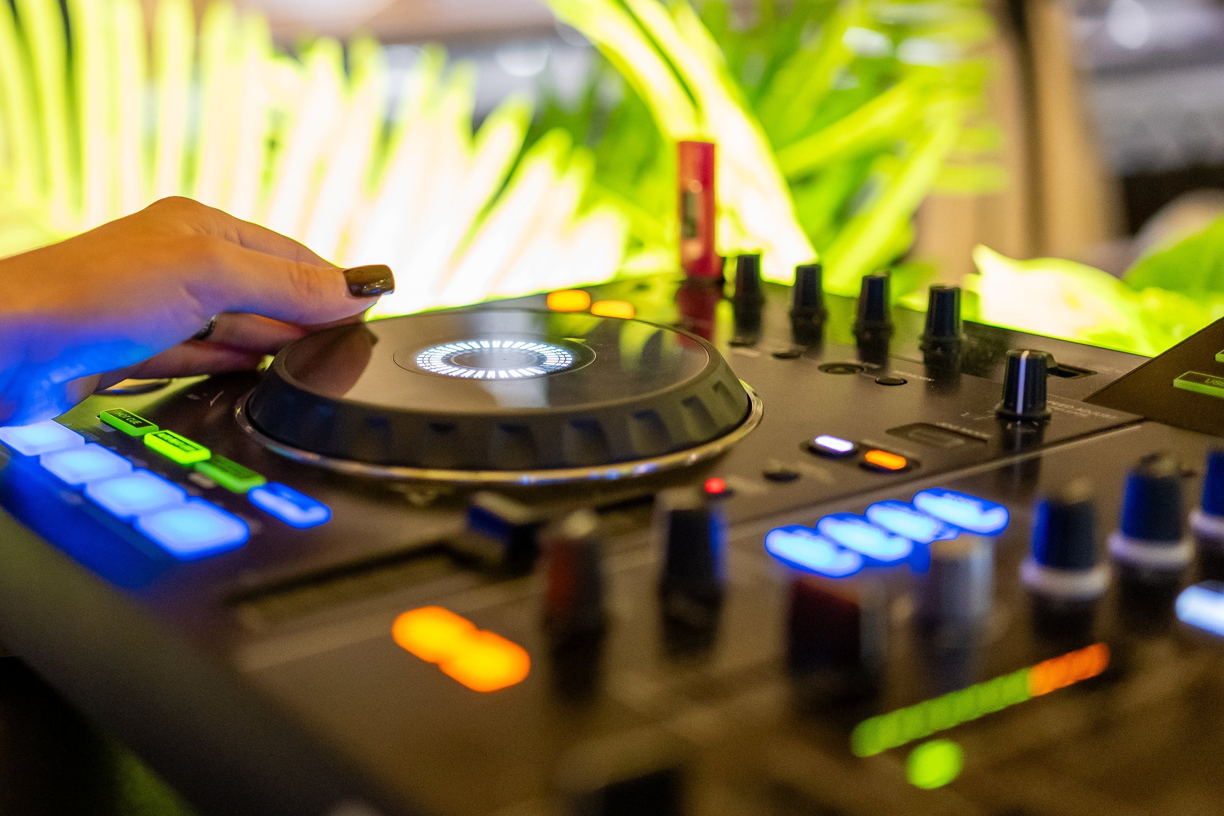 DJ adjusting turntable on professional mixer with glowing buttons, close-up of hand during vibrant party with green plants in background, modern music and nightlife concept.