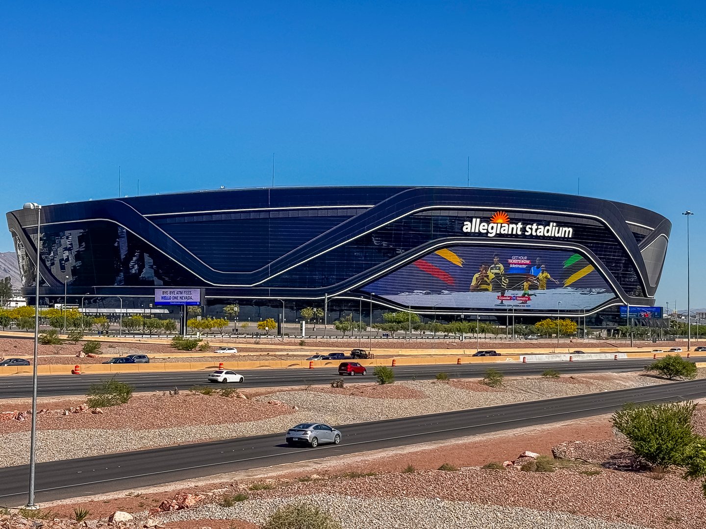 Las Vegas, NV, USA - May 12, 2024: Black Allegiant Stadium behind interstate 15 with international soccer sign under blue sky. Empty parking, cars on road, and green foliage