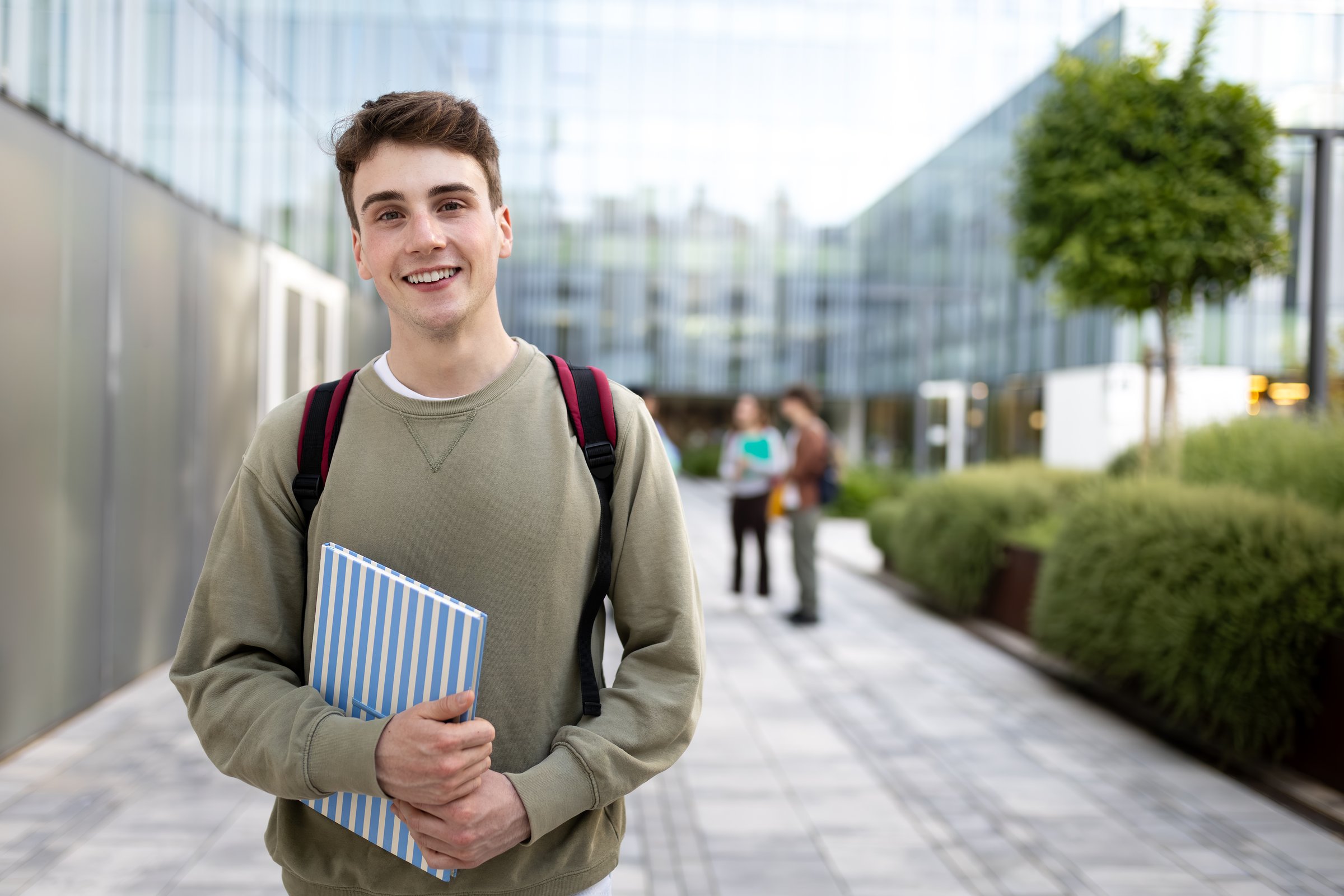Portrait of a smiling male student holding a book and wearing a backpack on a university campus