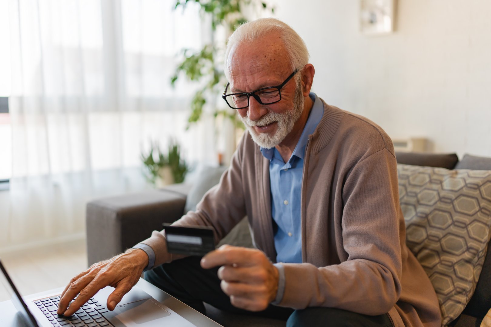 A cheerful elderly man with a beard sits comfortably on a sofa in a bright, naturally lit room. He uses a laptop while holding a credit card, smiling as he shop online or makes a secure digital payment