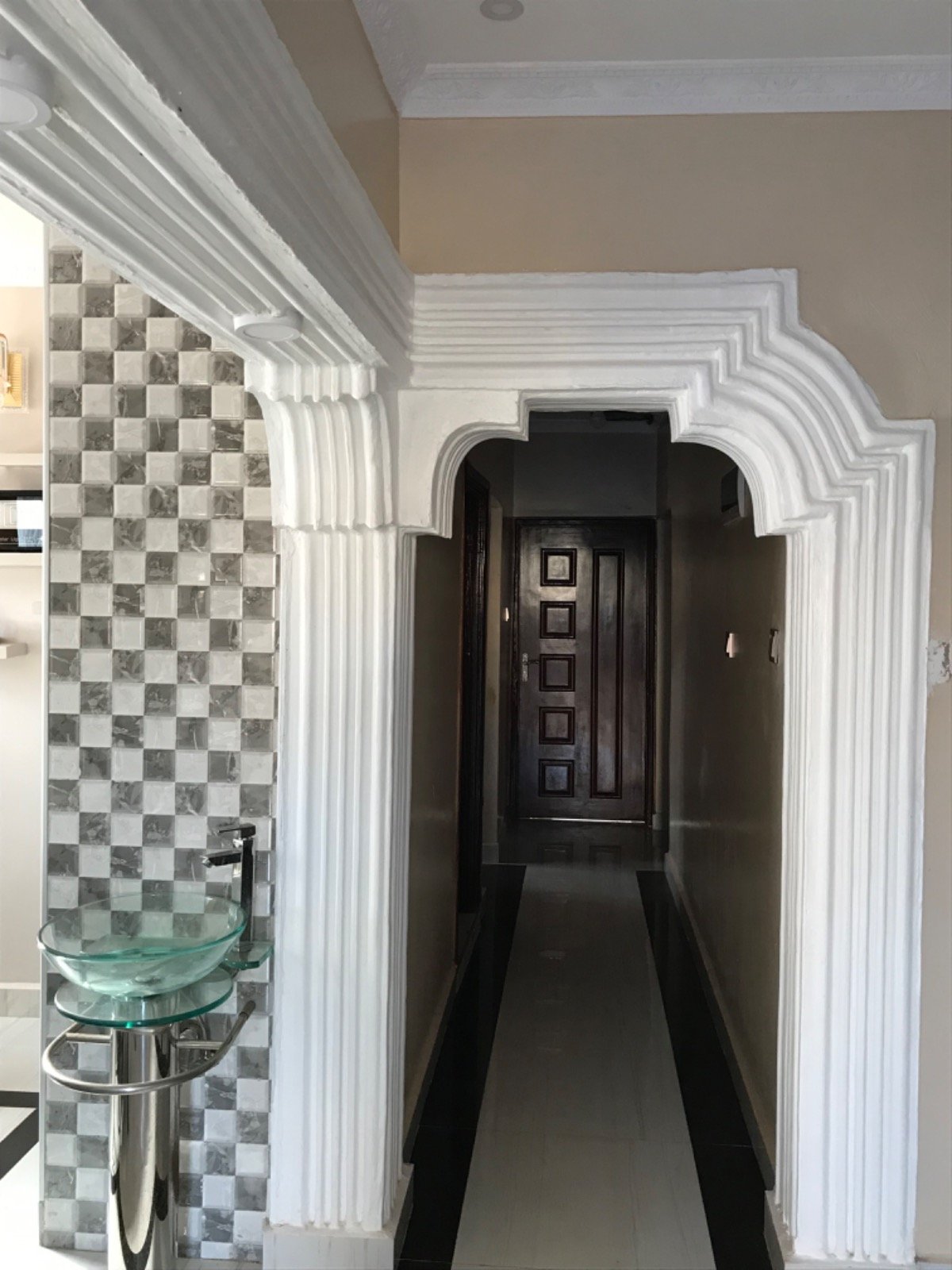 Interior archway with ornate white molding, leading to a hallway. Glass basin and tiled wall visible on the left.