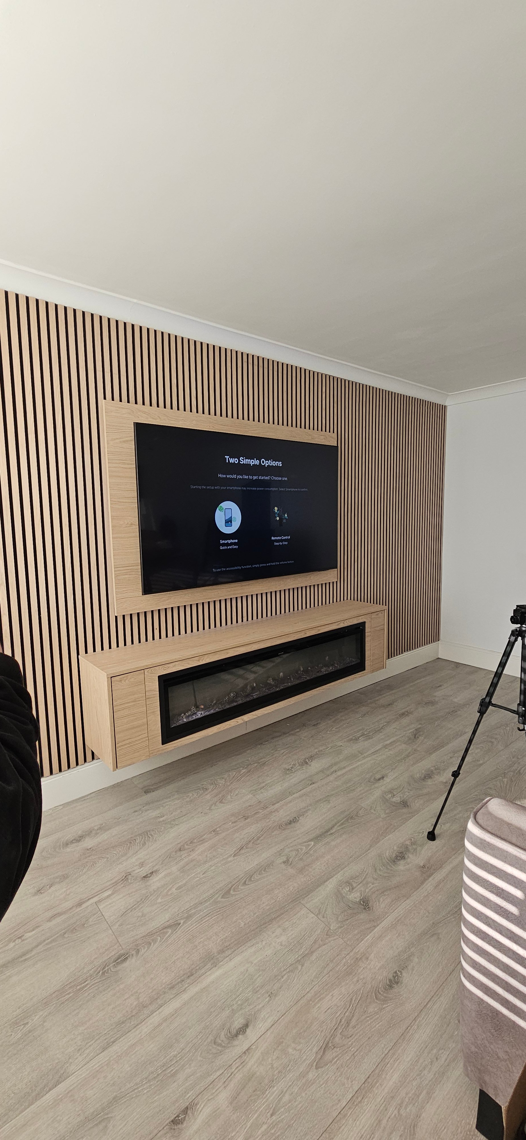 Modern living room with TV on a slatted wood accent wall, mounted above a fireplace, and tripod standing nearby.