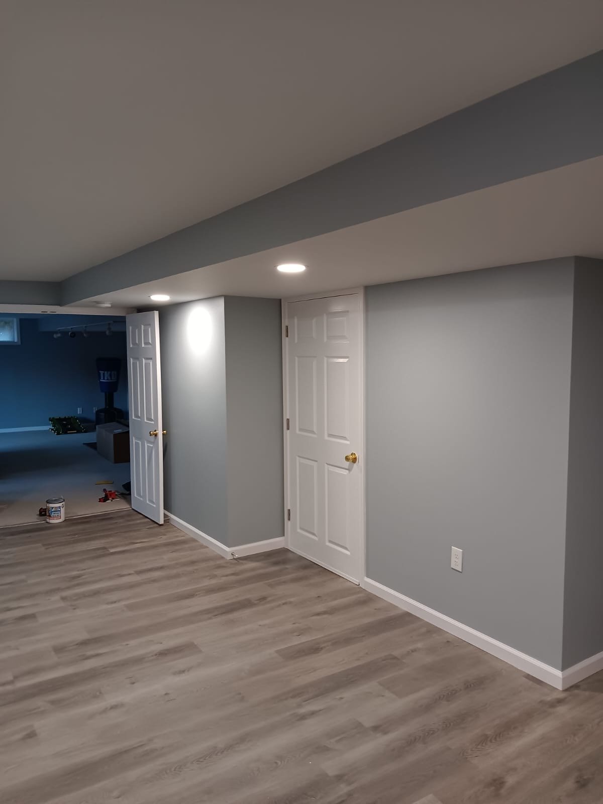 Finished basement with gray walls, wood flooring, recessed lights, and two white doors. Paint can visible on the floor.