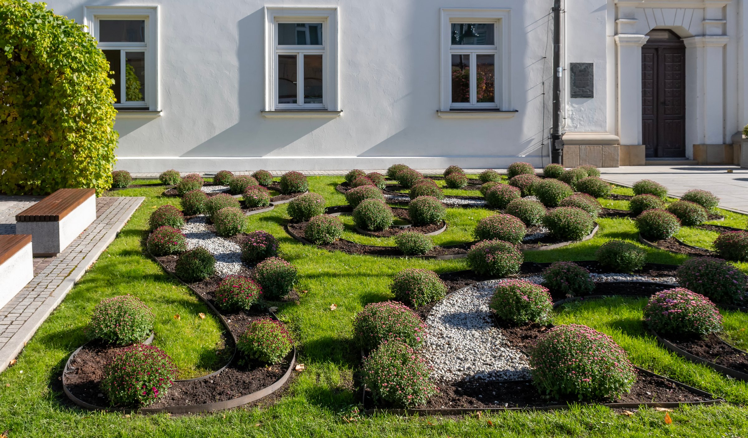 A carefully arranged garden featuring rounded shrubs, curved gravel strips, and a well-kept green lawn beside a bright building facade.