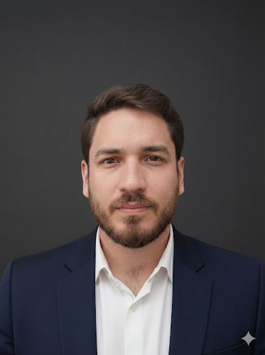 A man in a navy blazer and white shirt standing outdoors with greenery in the background.