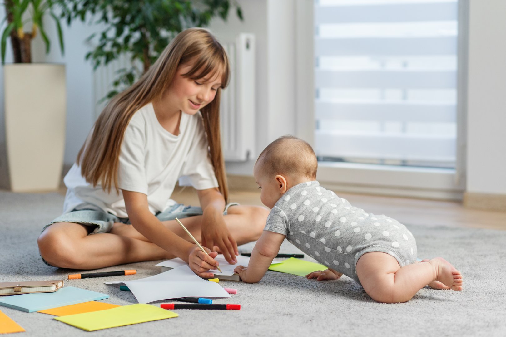 Older sister draws on paper with colored pens while baby in polka dot onesie curiously watches beside her on a soft carpet.