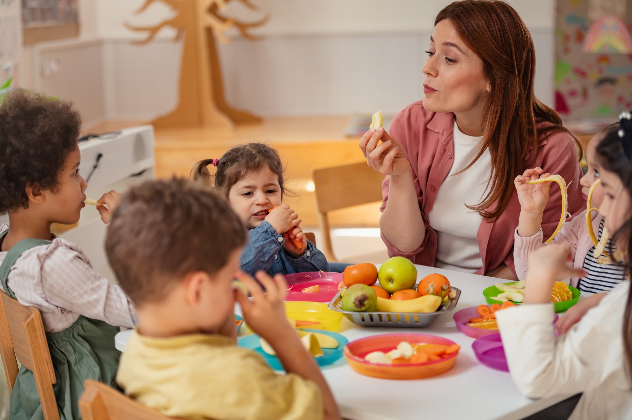 A preschool teacher smiles warmly as she interacts with children enjoying a healthy snack at a table. The children are focused on eating fruits and vegetables in a bright, cheerful classroom setting.