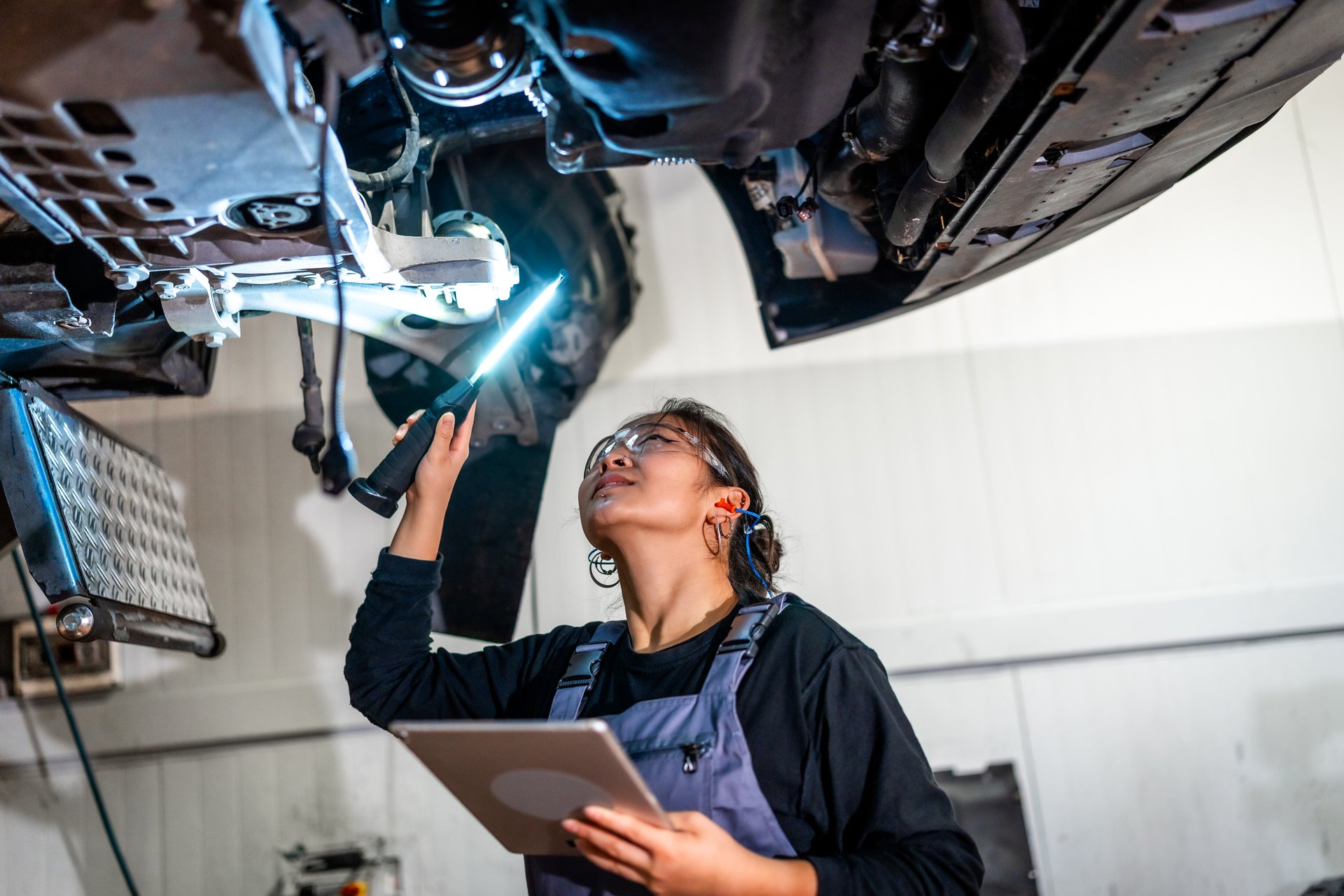 Young female mechanic using flashlight and digital tablet, inspecting underside of car raised on lift in auto repair shop