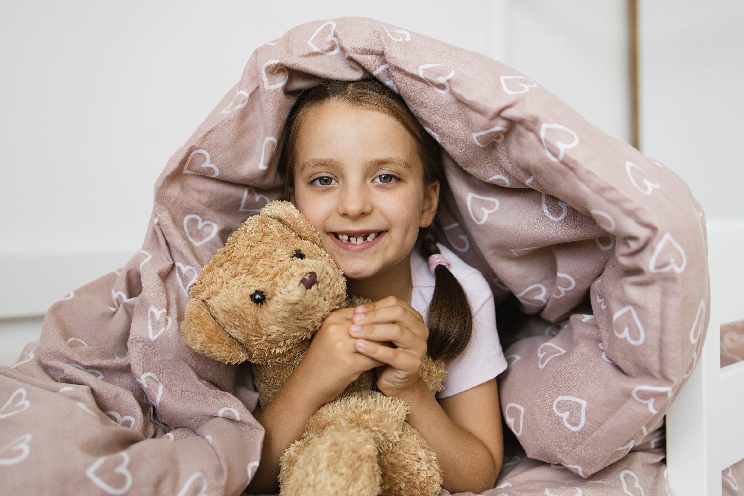 Little Caucasian girl with missing teeth smiles while lying under blanket holding teddy bear. Brown heart-patterned quilt adds warmth and comfort. Happy childhood moment captured in bedroom setting.