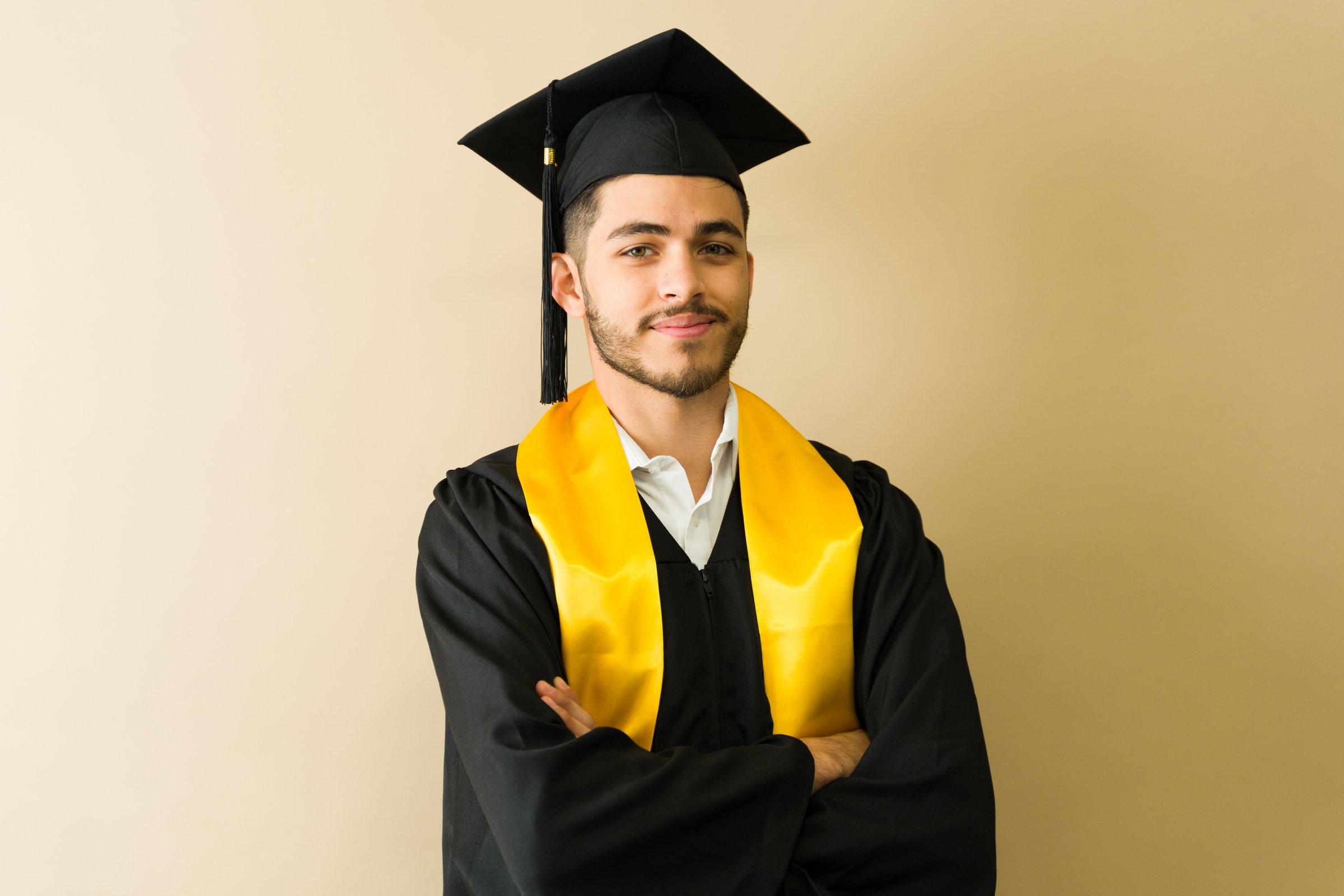 Proud graduate wearing a black gown and yellow sash, smiling with crossed arms after the graduation ceremony