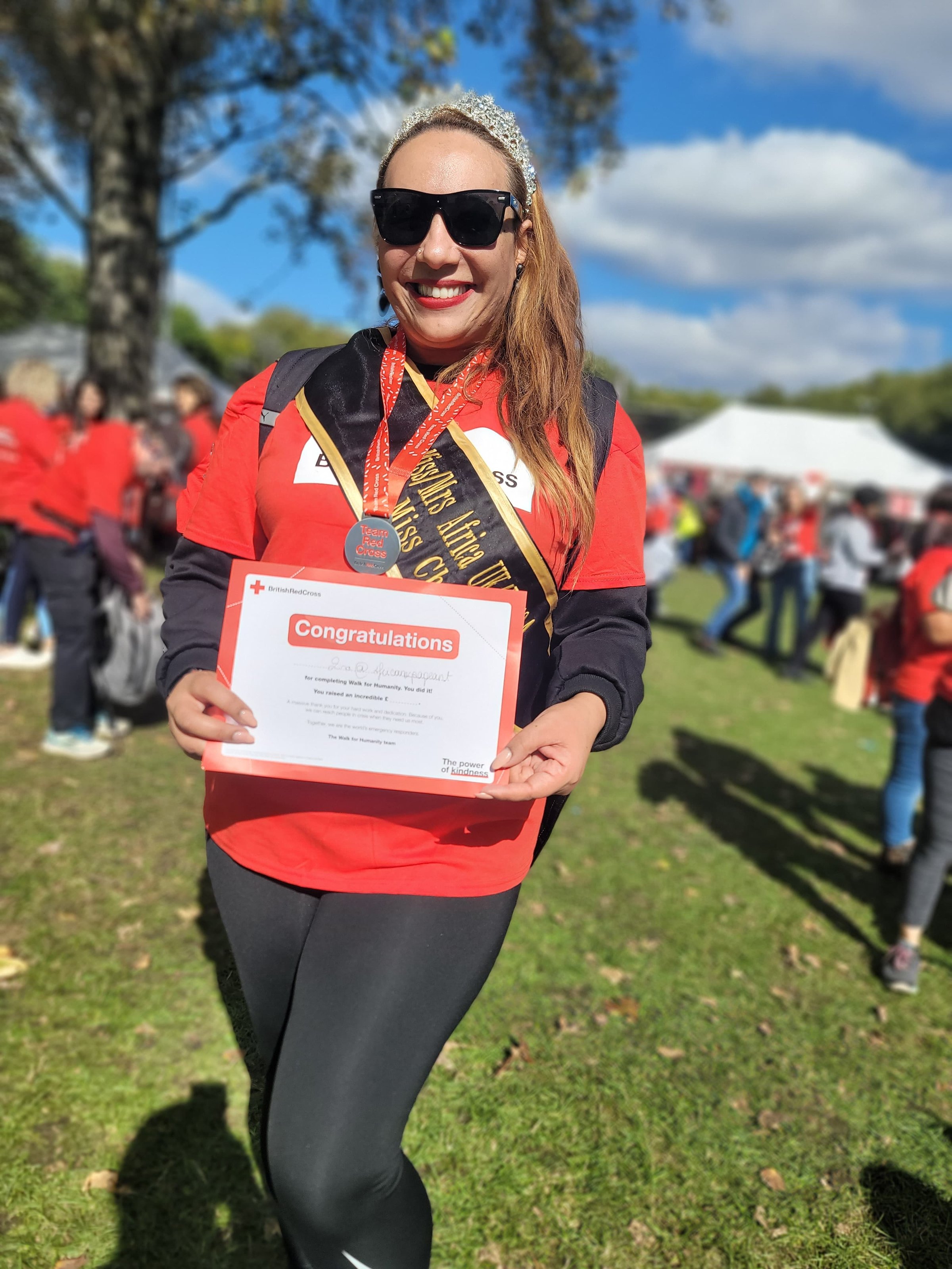 A woman wearing a tiara and sunglasses holds a certificate and smiles at an outdoor event.