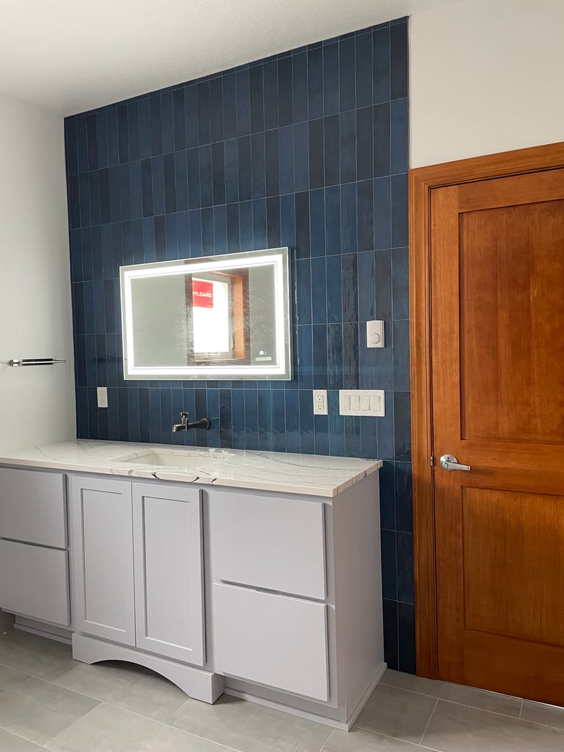 Modern bathroom with a white vanity, blue tiled wall, illuminated mirror, and wooden door.