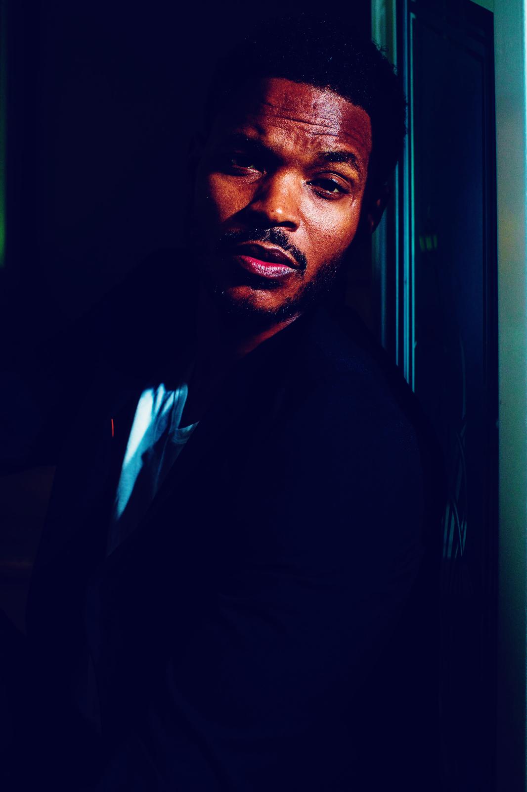 A man in a dark room, wearing a black jacket over a white shirt, looking towards the camera with a serious expression.