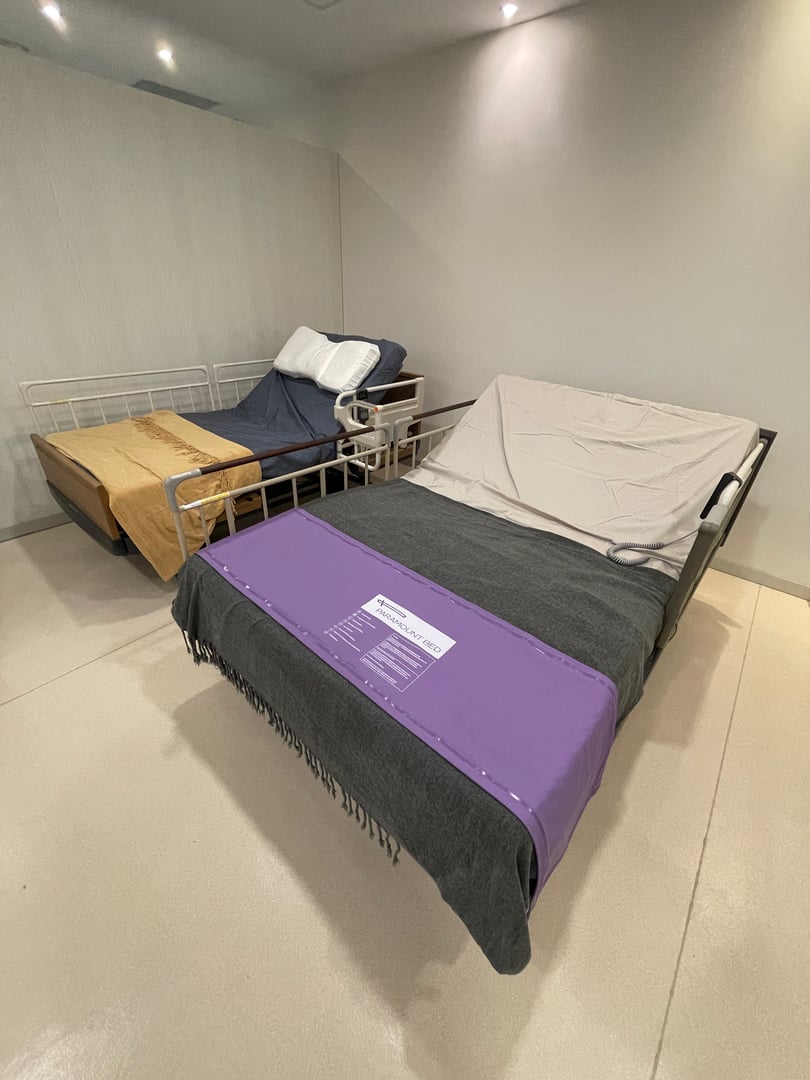 Two hospital-style beds with adjustable frames, one covered with a purple and gray blanket, in a plain room.