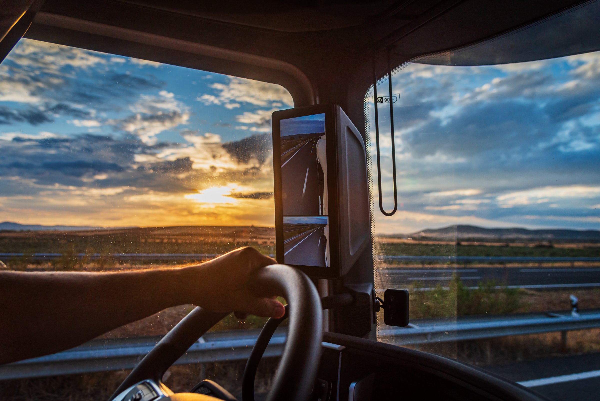 Truck cabin view with driver's hand on the wheel under a colorful sunset sky