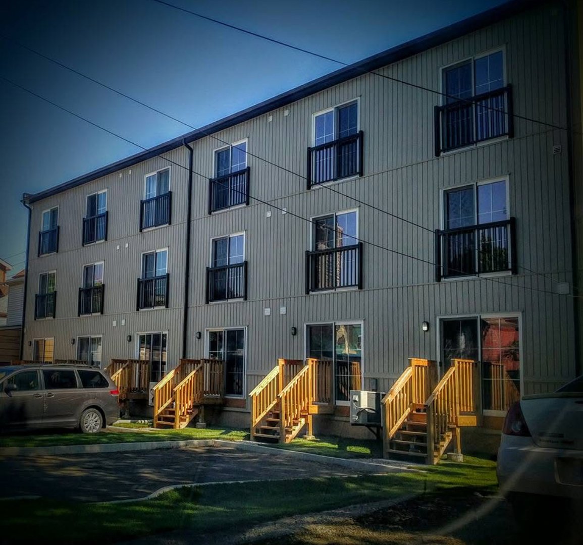 Three-story apartment building with wooden stairs, parking, and vehicles in front, under a clear blue sky.