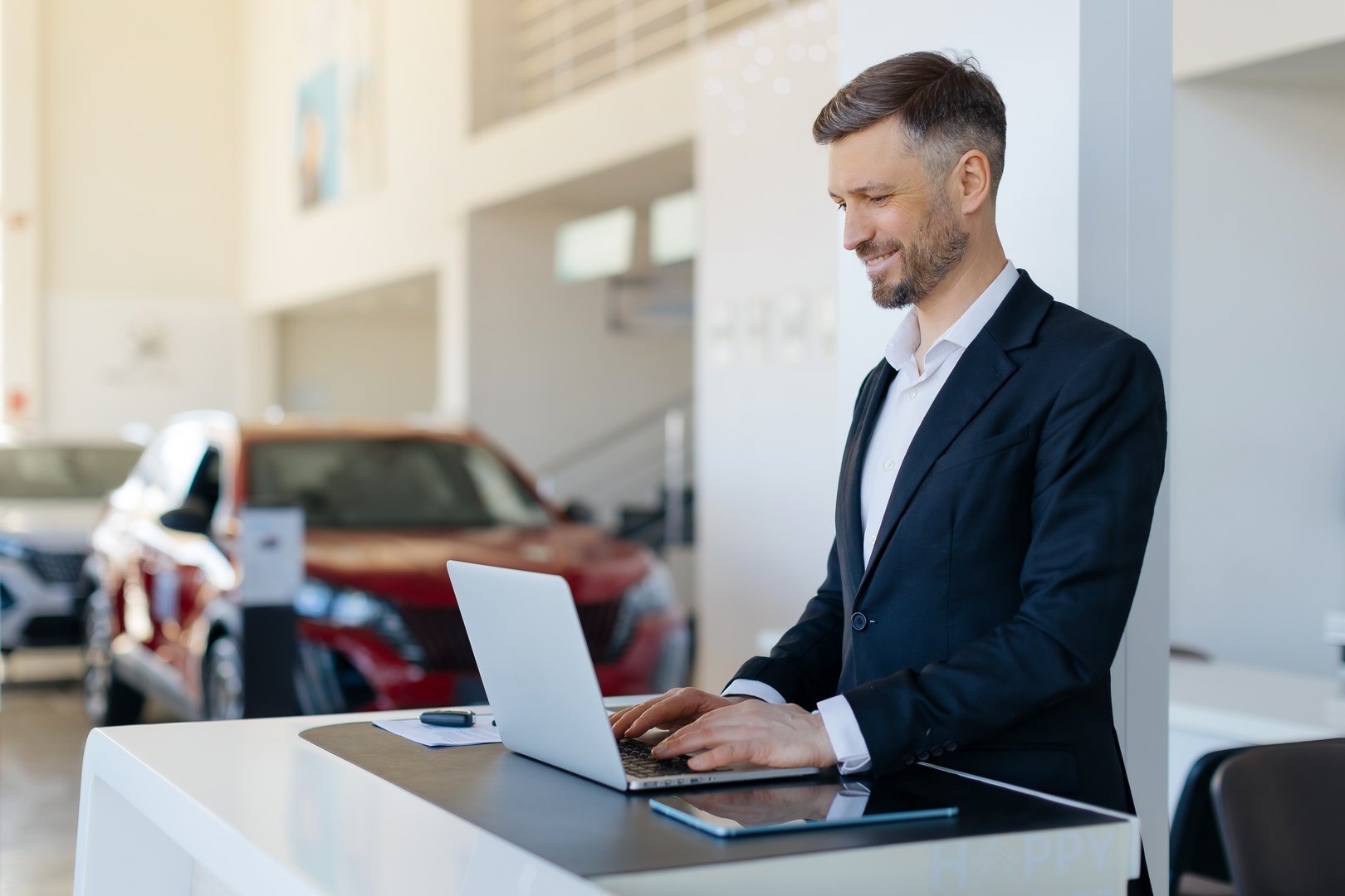 Positive handsome car salesman using laptop computer at work desk in auto dealership center. Cheerful millennial automobile manager working with pc in showroom store, free space