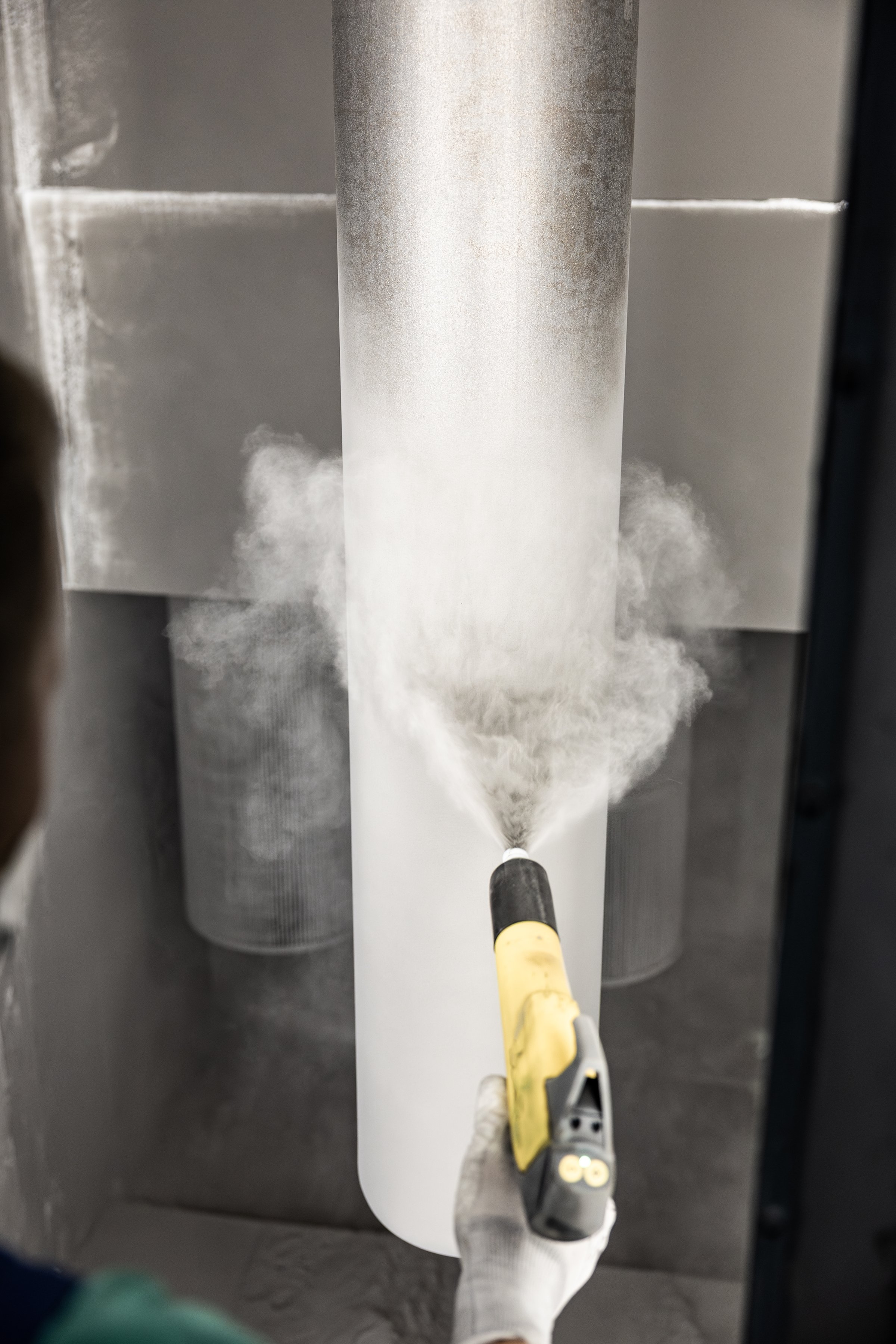 A technician uses a spray gun to apply white powder coating to a large vertical metal gas cylinder in an enclosed booth. No visible logos or branding