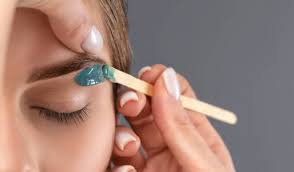 Close-up of a person applying green wax to shape eyebrows using a wooden spatula.