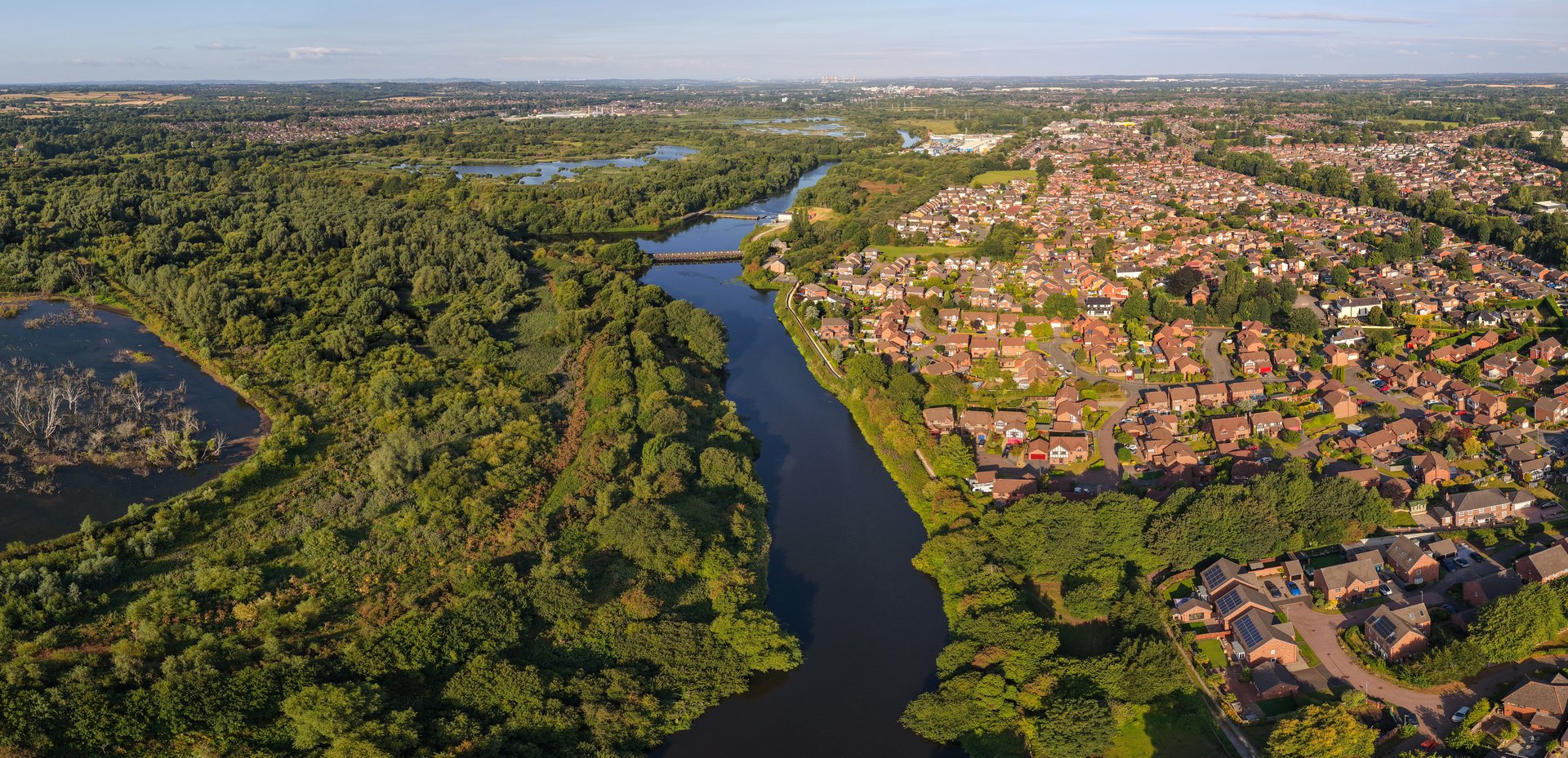 Aerial panoramic view of Woolston, near Warrington, with the River Mersey winding past residential areas, dense woodlands, and a wetlands