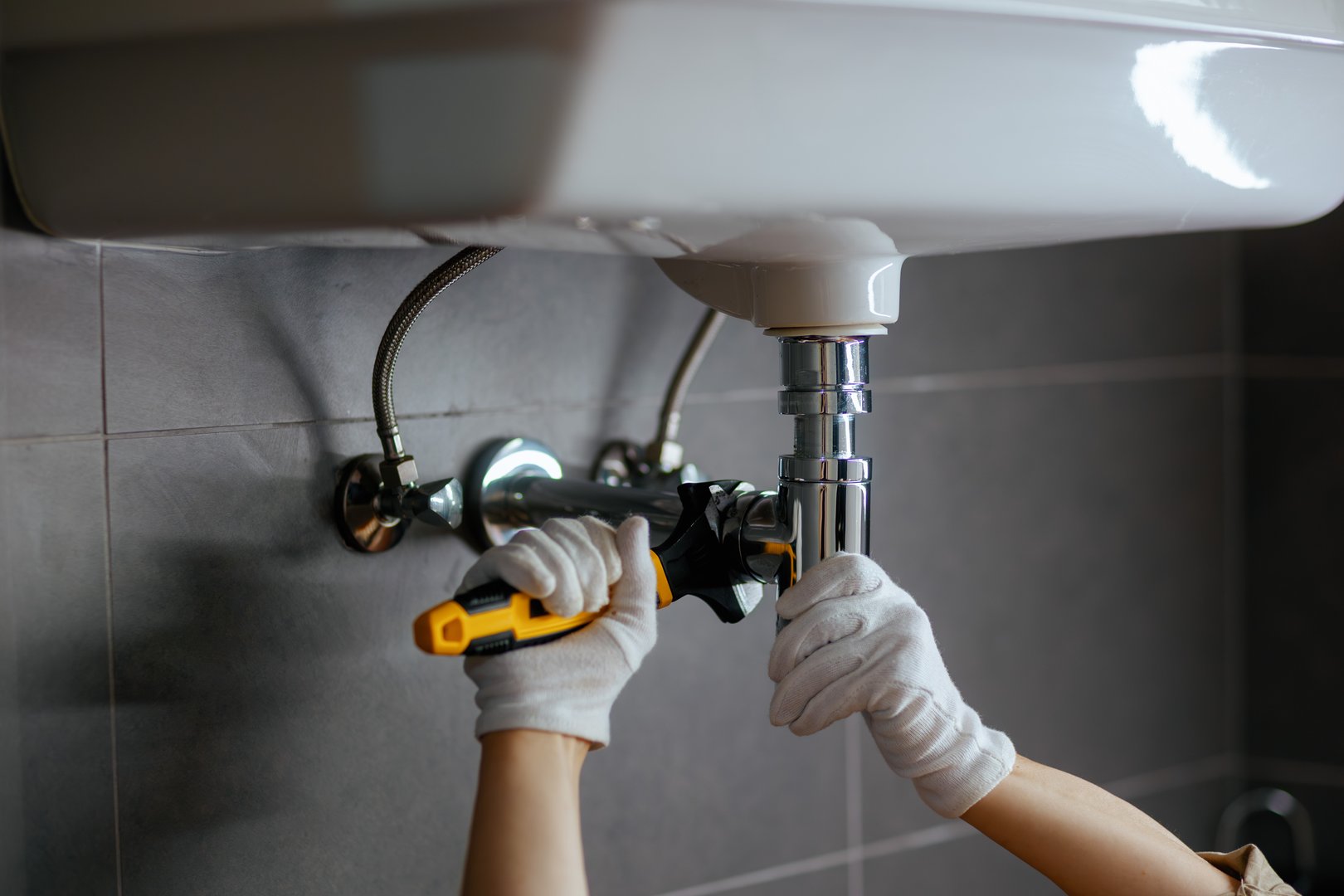 A plumber tightens a chrome drain pipe under a white sink with a wrench, focusing on a home repair task in a bathroom setting.