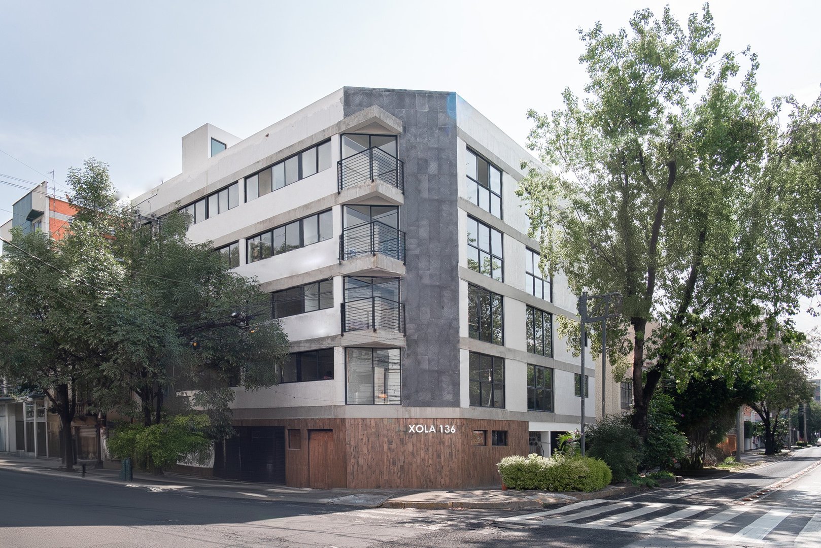 Modern five-story building with large windows on a street corner, surrounded by trees.